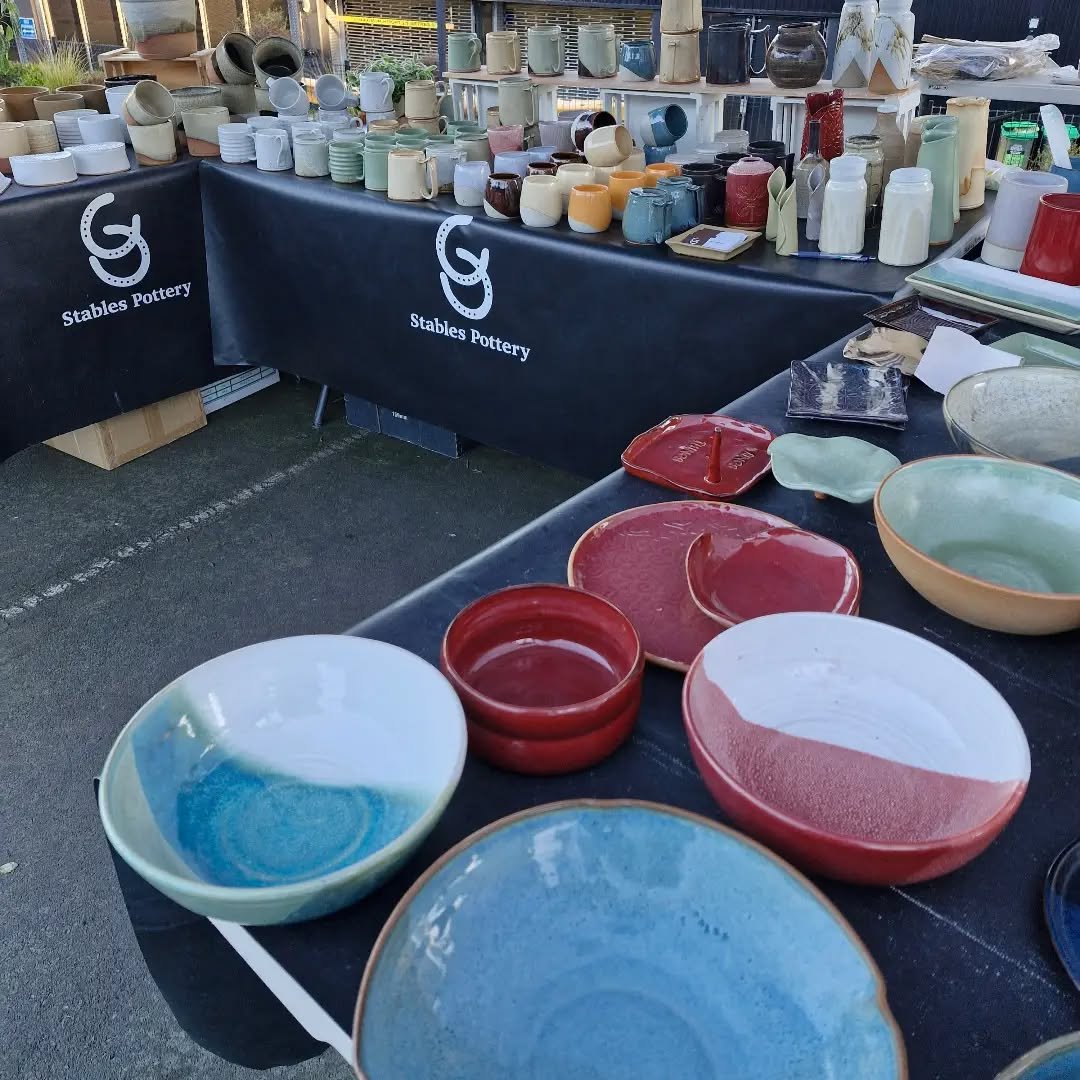 A display of handcrafted pottery including bowls, cups, and vases with various glazes and textures, arranged on a black tray underneath a large green monstera leaf.