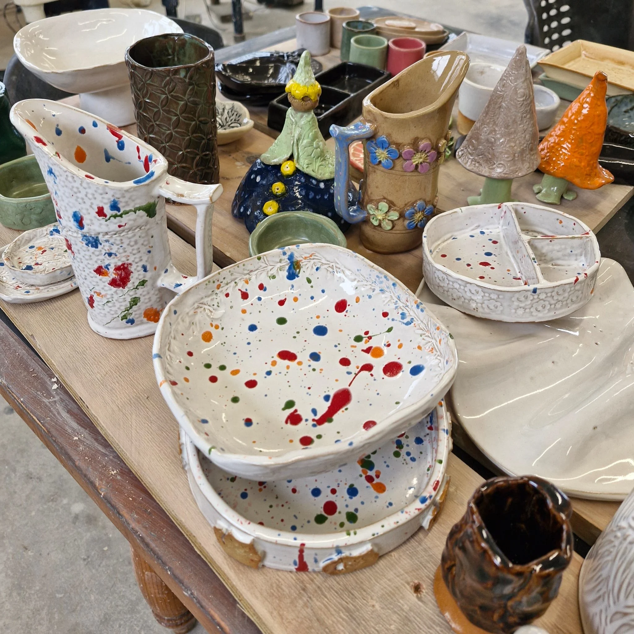 Various handmade ceramic pottery pieces, including cups, bowls, and decorative items, displayed on a wooden table. The pieces are colorful with splatter and floral patterns.