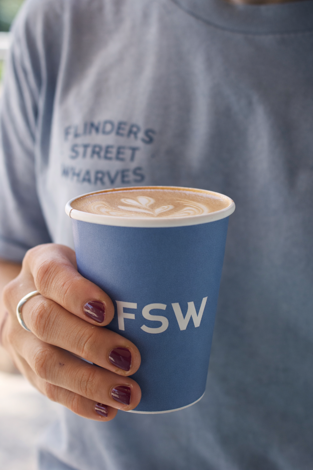 A person holding a blue paper cup with white lettering that says 'FSW' and a latte with latte art on top, wearing a gray t-shirt with blue text that says 'FLINDERS STREET WHARVES.'