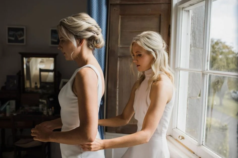 A woman in a white dress helps another woman in a white dress adjust her clothing by a window in a stately home with wooden walls. Brocket Hall.