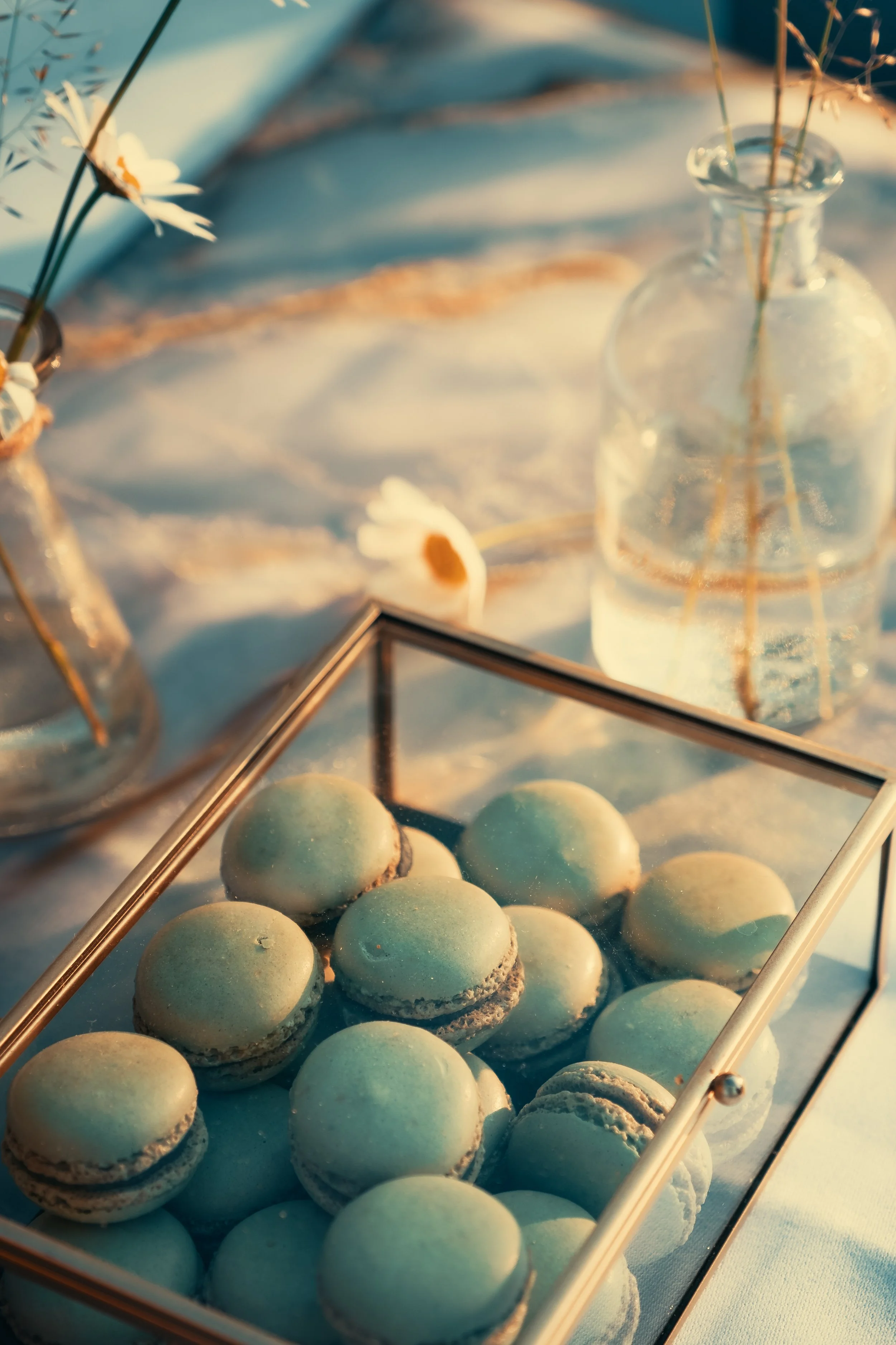 Groene macarons in een glazen doos met een glazen flesje met bloemen op de achtergrond, bij warm licht.