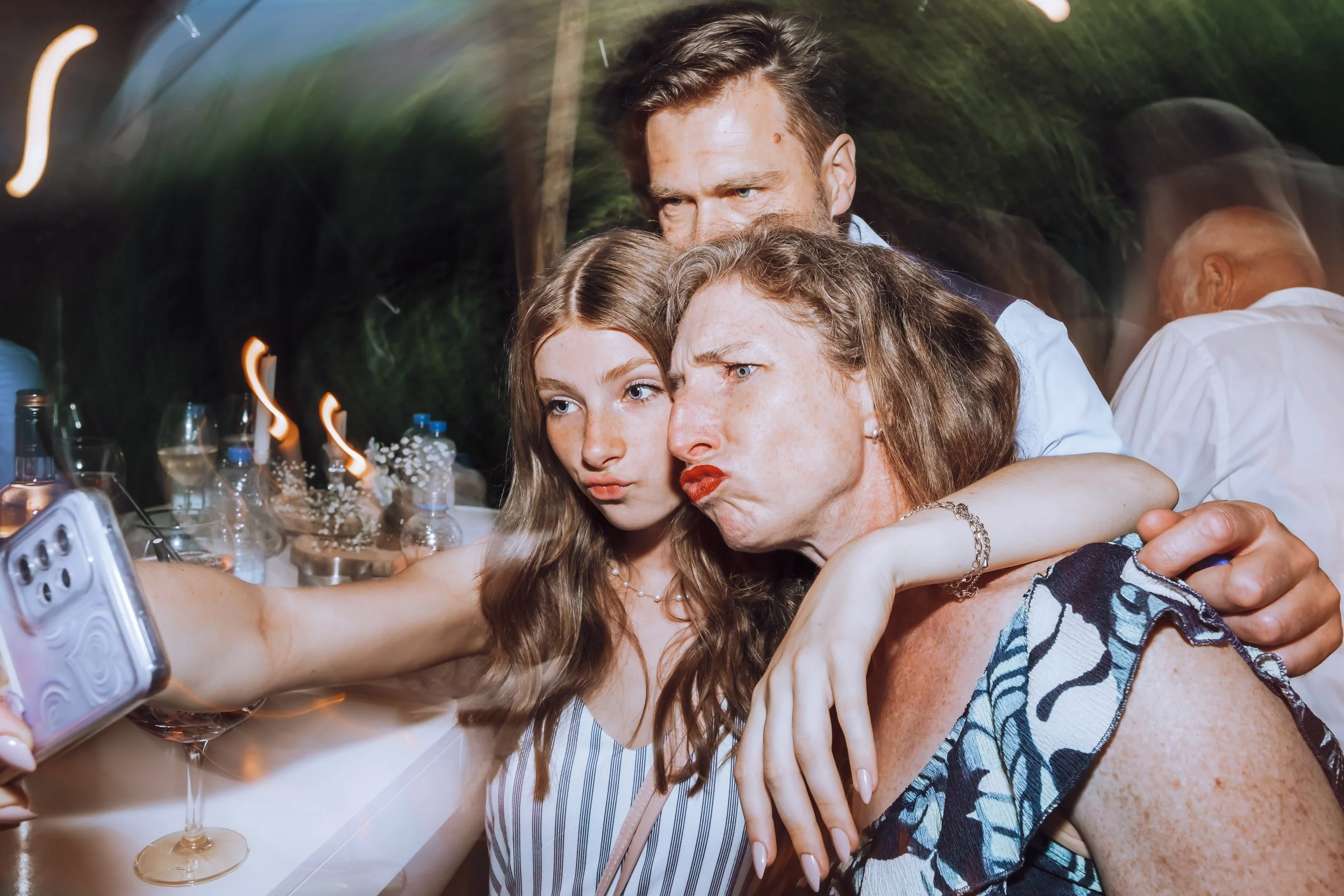 Vijf mensen maken een groepsselfie aan een feesttafel, omringd door drankjes en glaasjes, in een buitenomgeving met donkergroene achtergrond.
