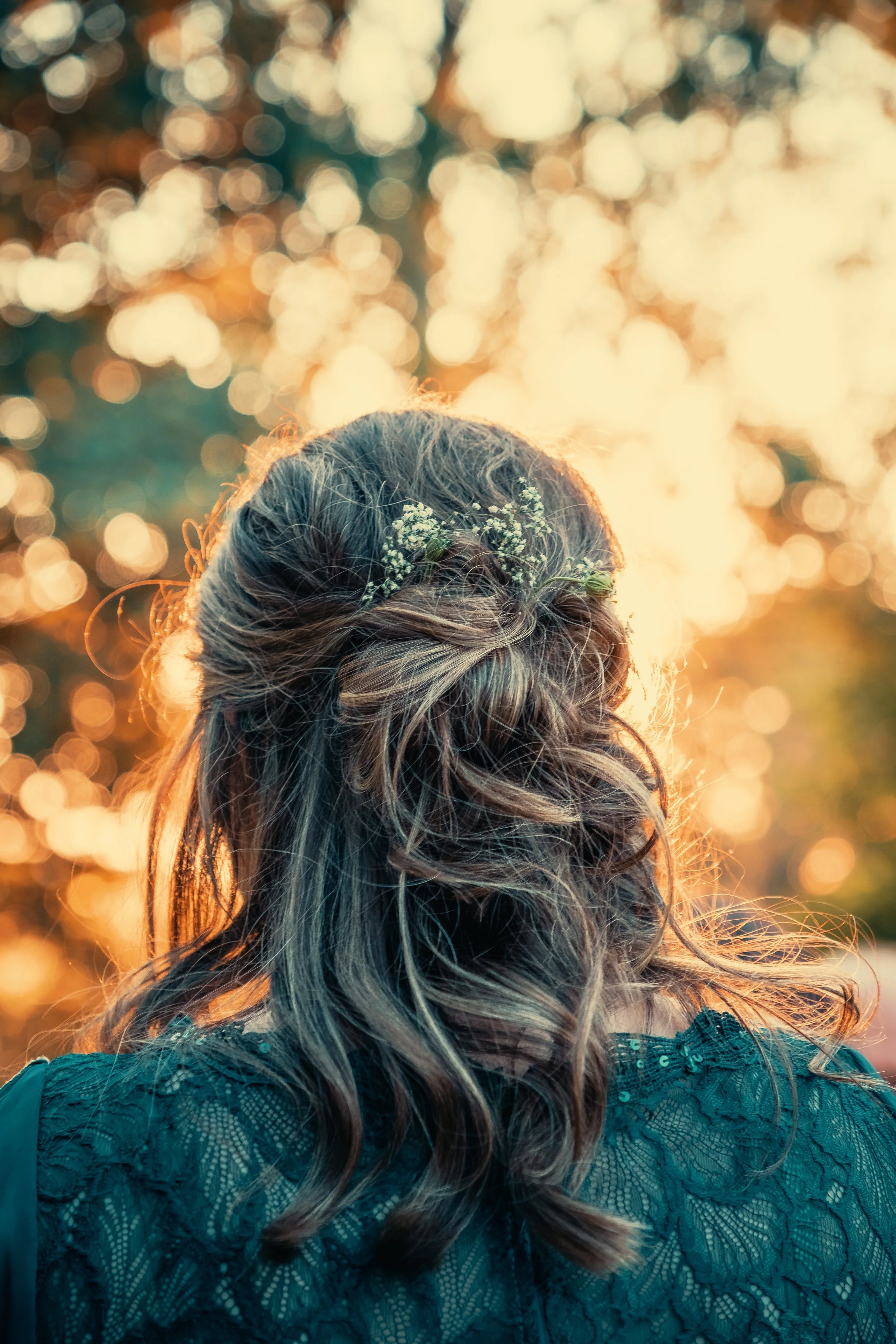 De achterkant van een vrouw met gewikkeld haar en een kleine bloem in haar haar, tijdens zonsondergang met een bokeh achtergrond van licht en bomen.
