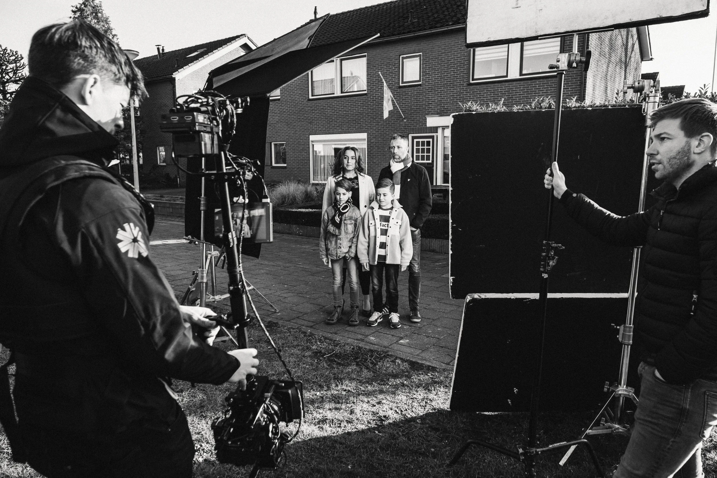 Mensen die filmen of fotograferen een gezin dat buiten voor een huis staat, met filmspullen en apparatuur op een filmset.