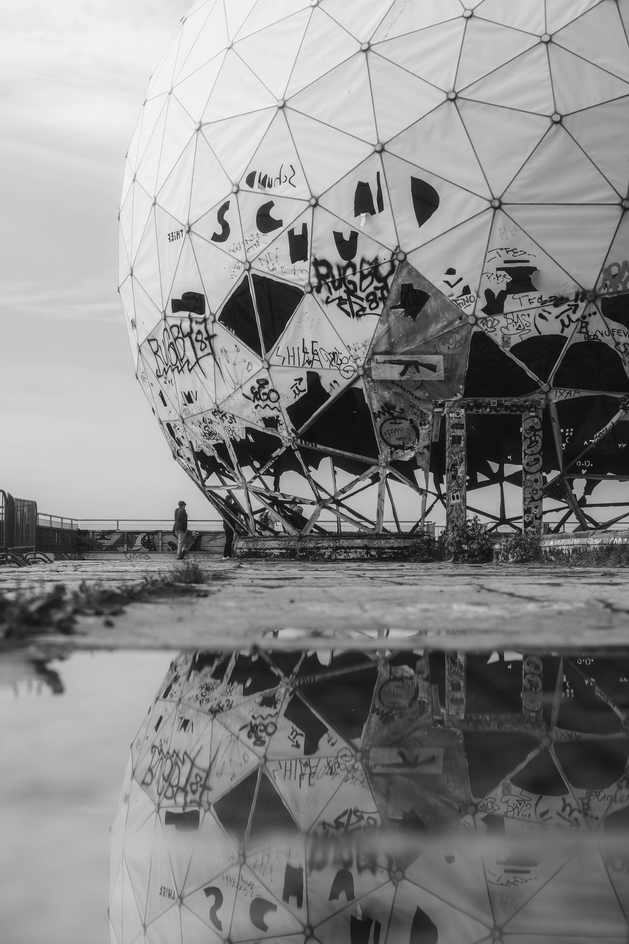 Een grote, metalen, gespiegelde structuur met graffiti en tags, met een weerspiegeling in een plas water op de grond onderaan de afbeelding.