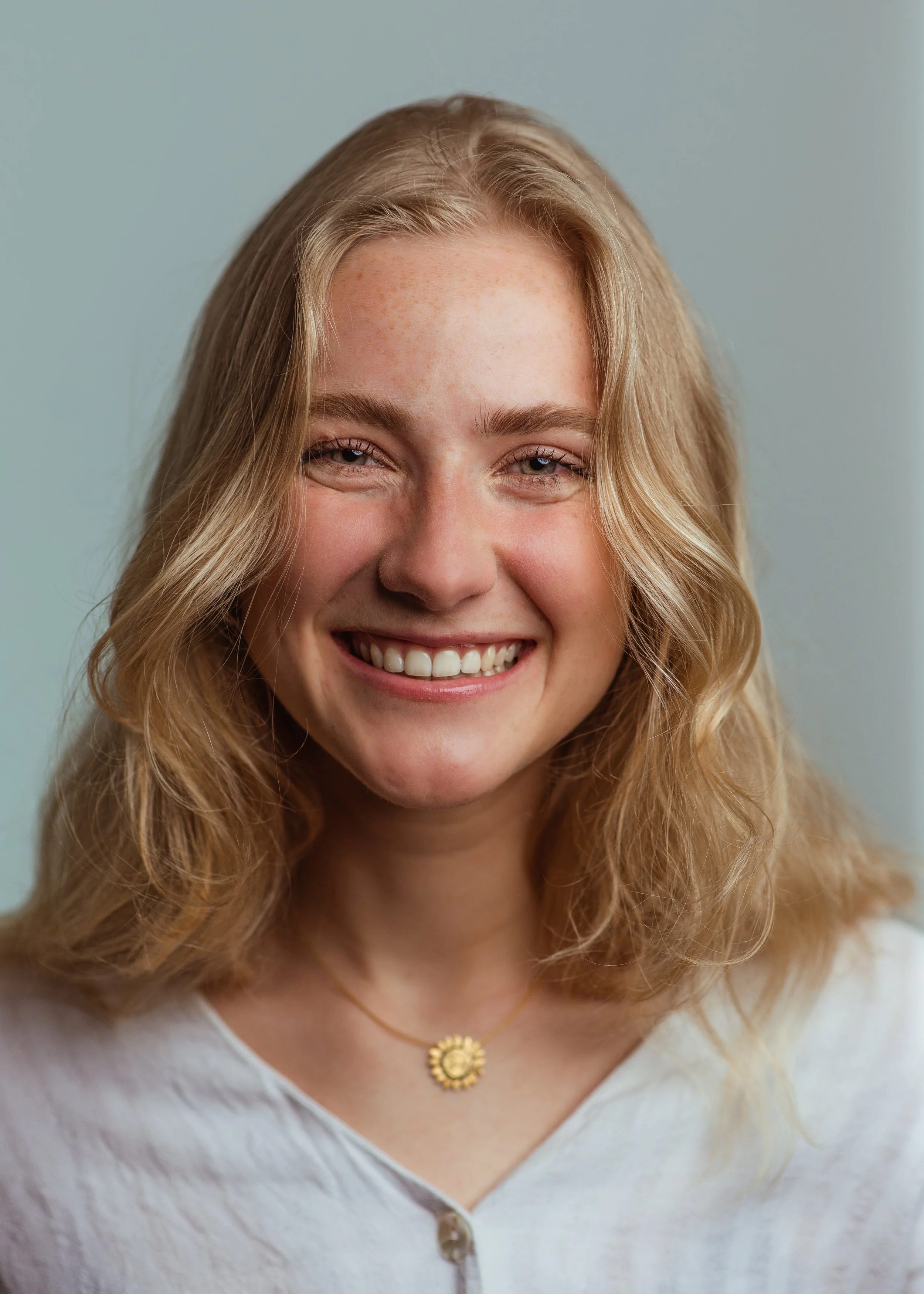 Close-up van een lachende jonge vrouw met lange, golvende blonde haren, witte tanden, en een gouden ketting met een bloemvormige hanger, gekleed in een witte top.