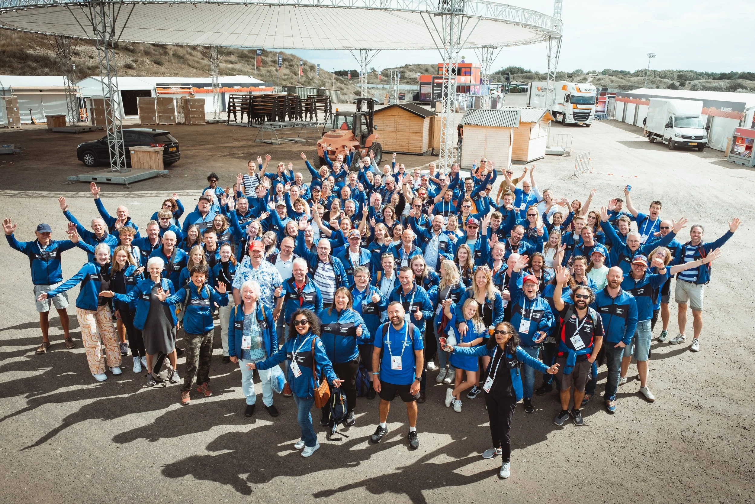 Grote groep mensen in blauwe jassen die samen poseert en zwaait bij een evenement op een open locatie met tijdelijke gebouwen en vrachtwagens op de achtergrond.