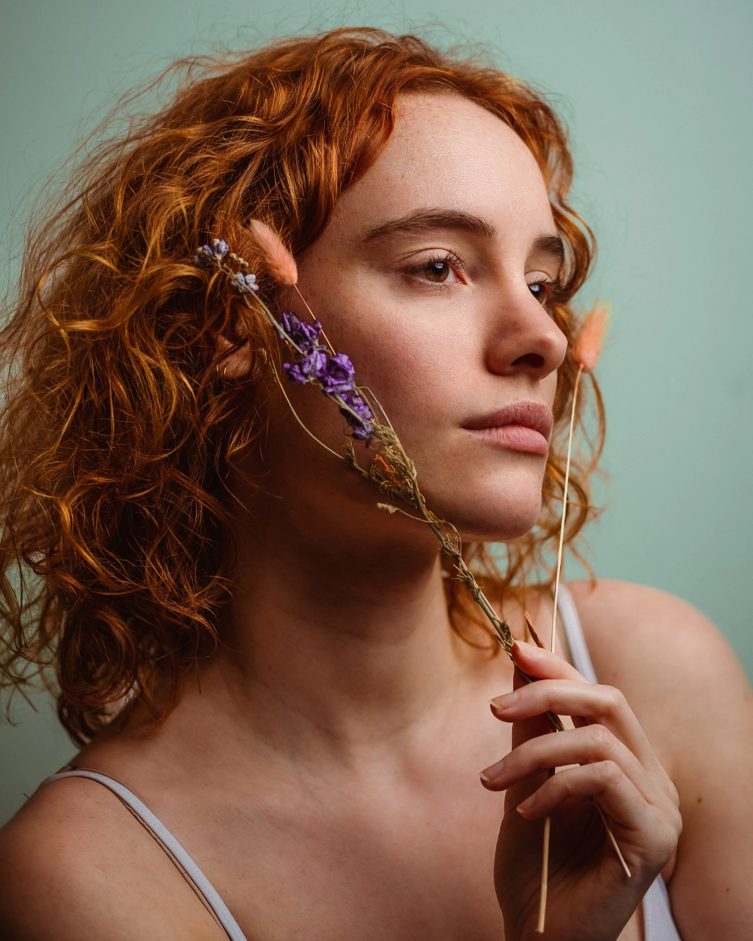 Close-up van een vrouw met krullend rood haar die een tak met paarse bloemen vasthoudt tijdens een fotoshoot, terwijl ze naar rechts kijkt.