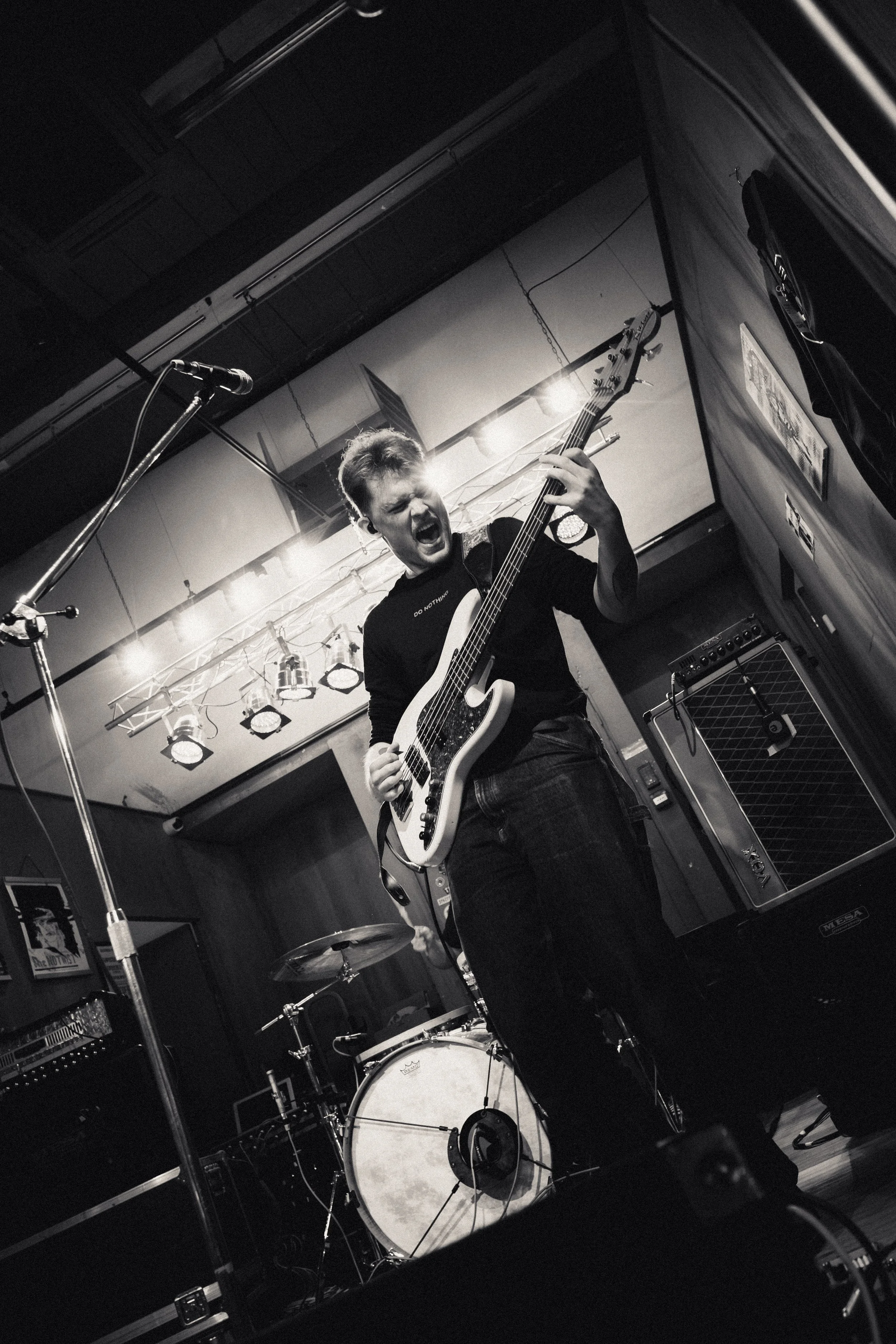 Een muzikant speelt gitaar en zingt krachtig op een podium, omgeven door muziekapparatuur en een drumstel.