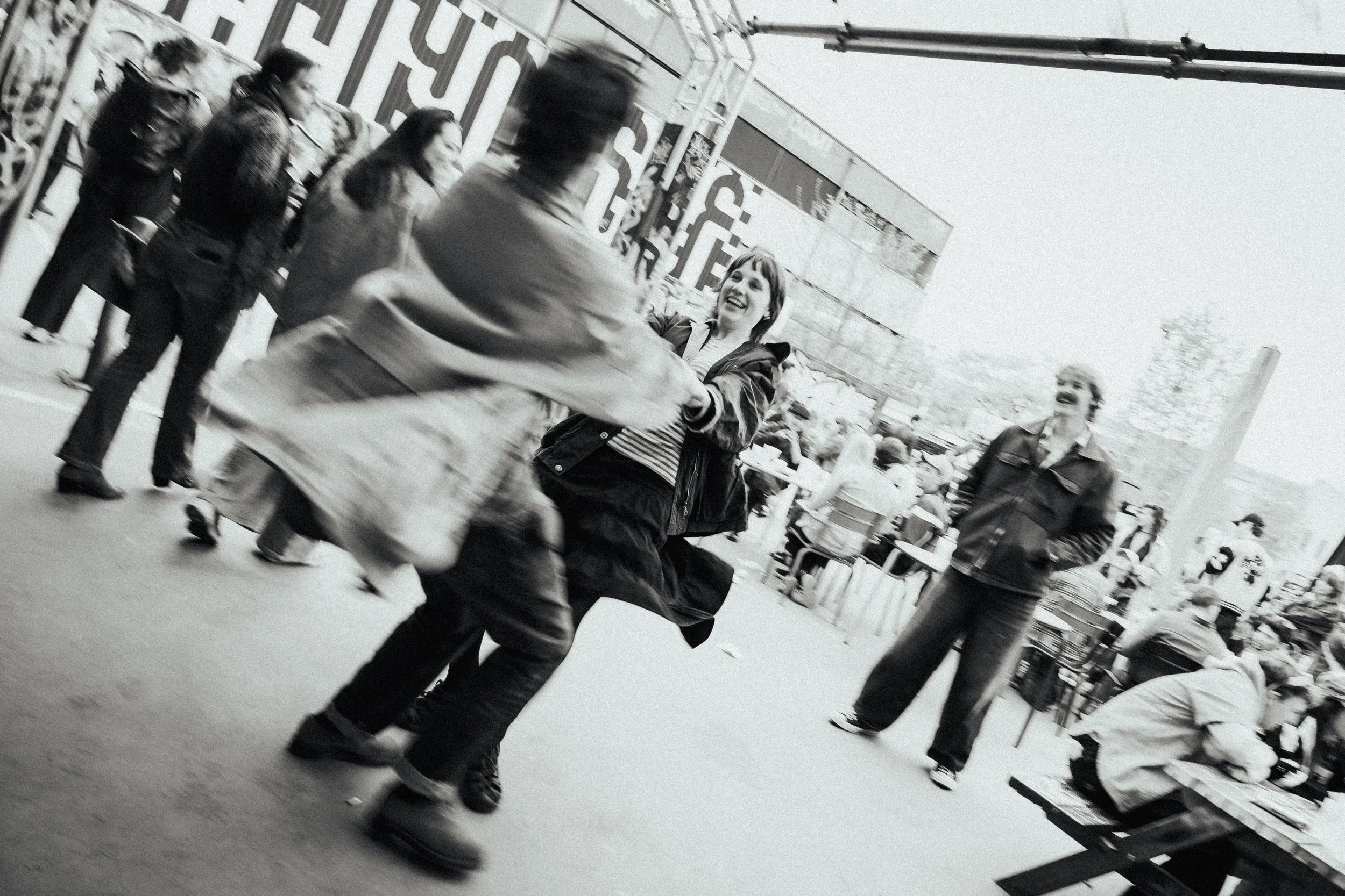 Mensen lopen over een markt in de openlucht, vanaf een afstand gezien. Een vrouw glimlacht en kijkt naar een man die haar een hand of object aanbiedt. Er zijn meerdere mensen op de achtergrond, en marktkramen en goederen zijn zichtbaar.