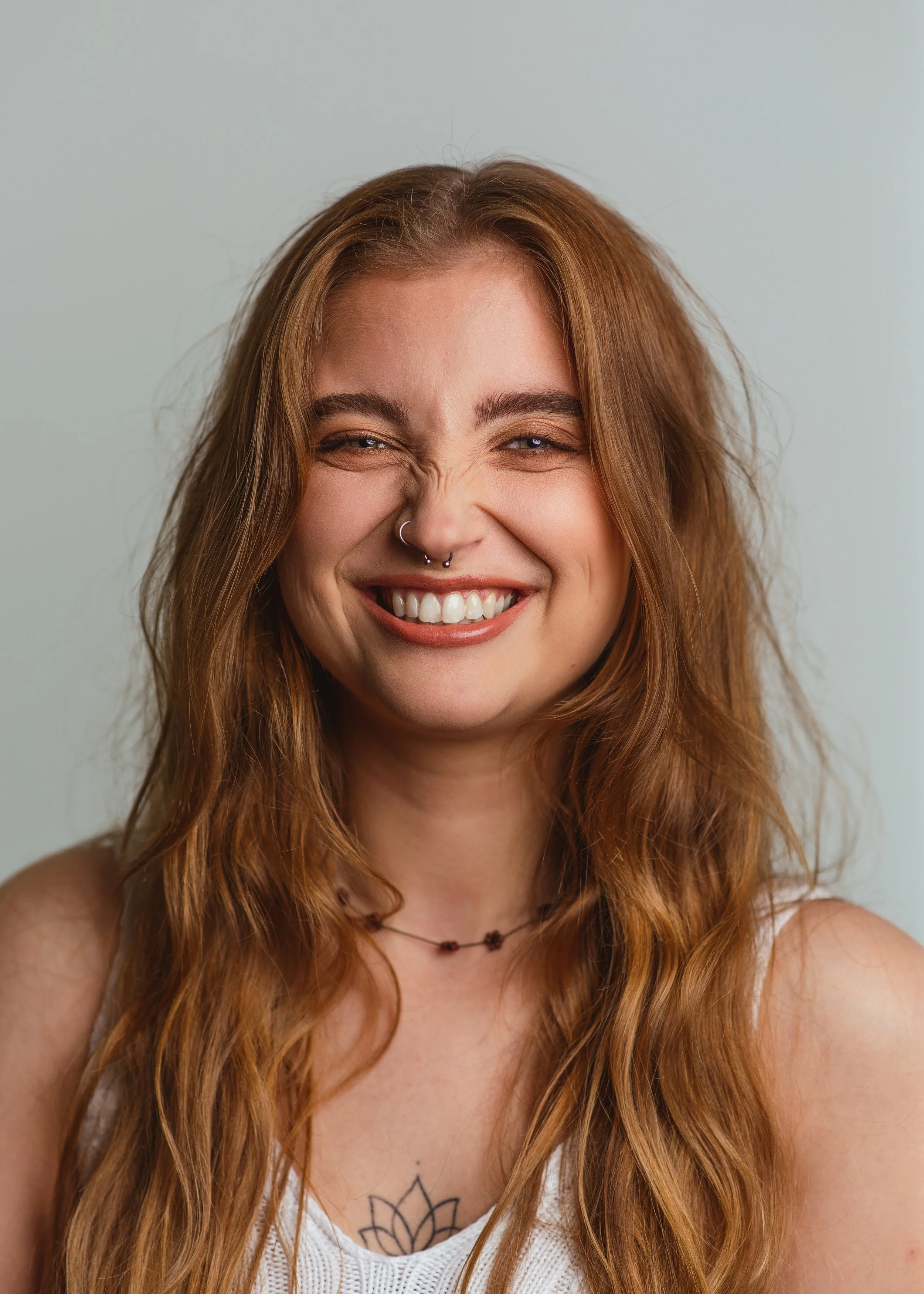 Close-up van een lachende jonge vrouw met lang, krullend, rood haar, een neuspiercing, een subtiele tatoeage op haar borst, en een witte top, voor een effen lichtgrijze achtergrond.
