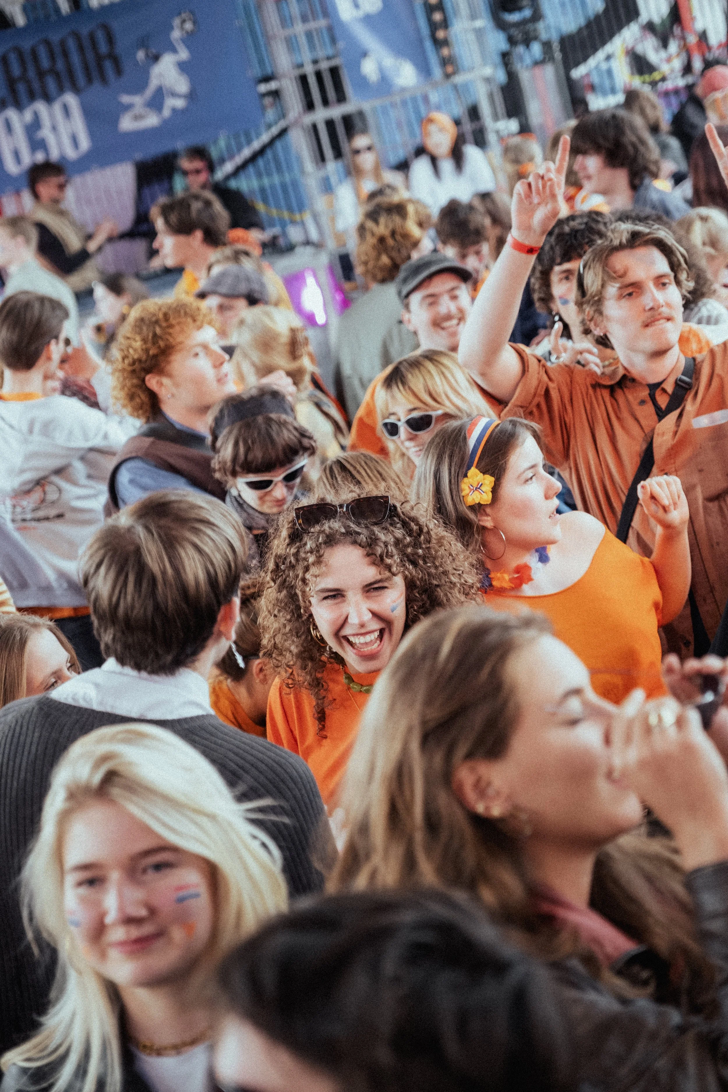 Een grote groep mensen die samen feest vieren, velen dragen oranje kleding en accessoires, en hebben gezichtsbeschilderingen in de kleuren van de Nederlandse vlag, wat wijst op een Nederlandse nationale feestdag.