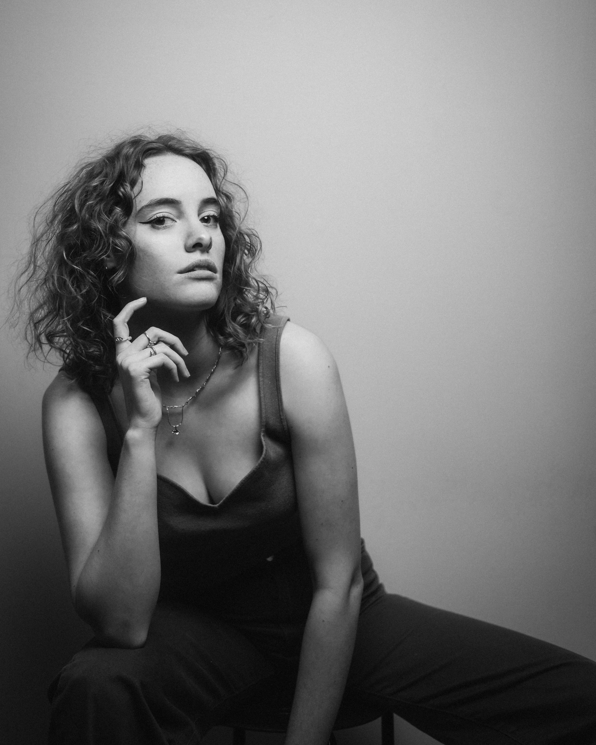Zwart-wit foto van een jonge vrouw met gekruld haar die naar links kijkt, haar hand onder haar kin, zittend op een bank met een effen muur op de achtergrond.