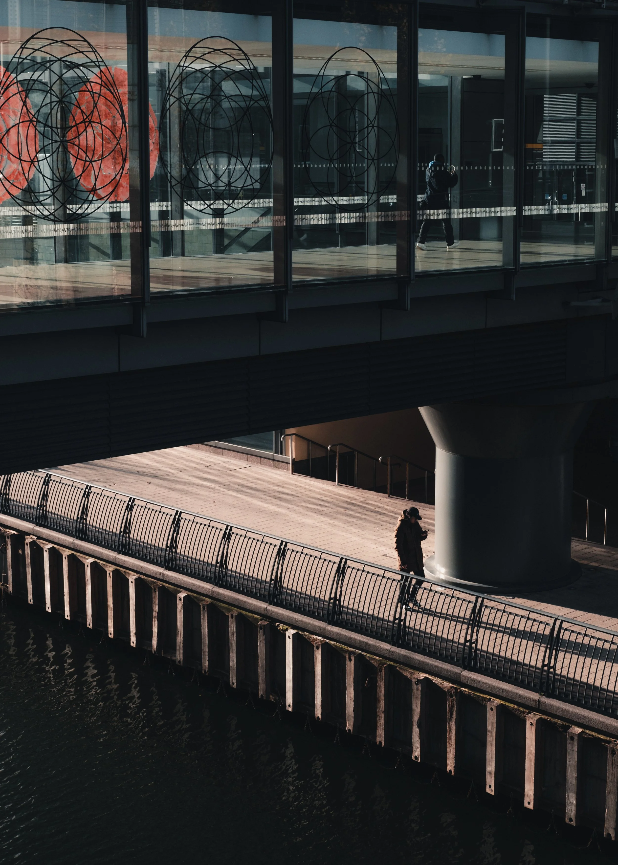 Een modern gebouw met grote glazen ramen en decoratieve geometrische patronen, gelegen aan een waterkant met een houten pier en een paar mensen die wandelen, waaronder een persoon met een hoed en een persoon die in een hoodie staat.