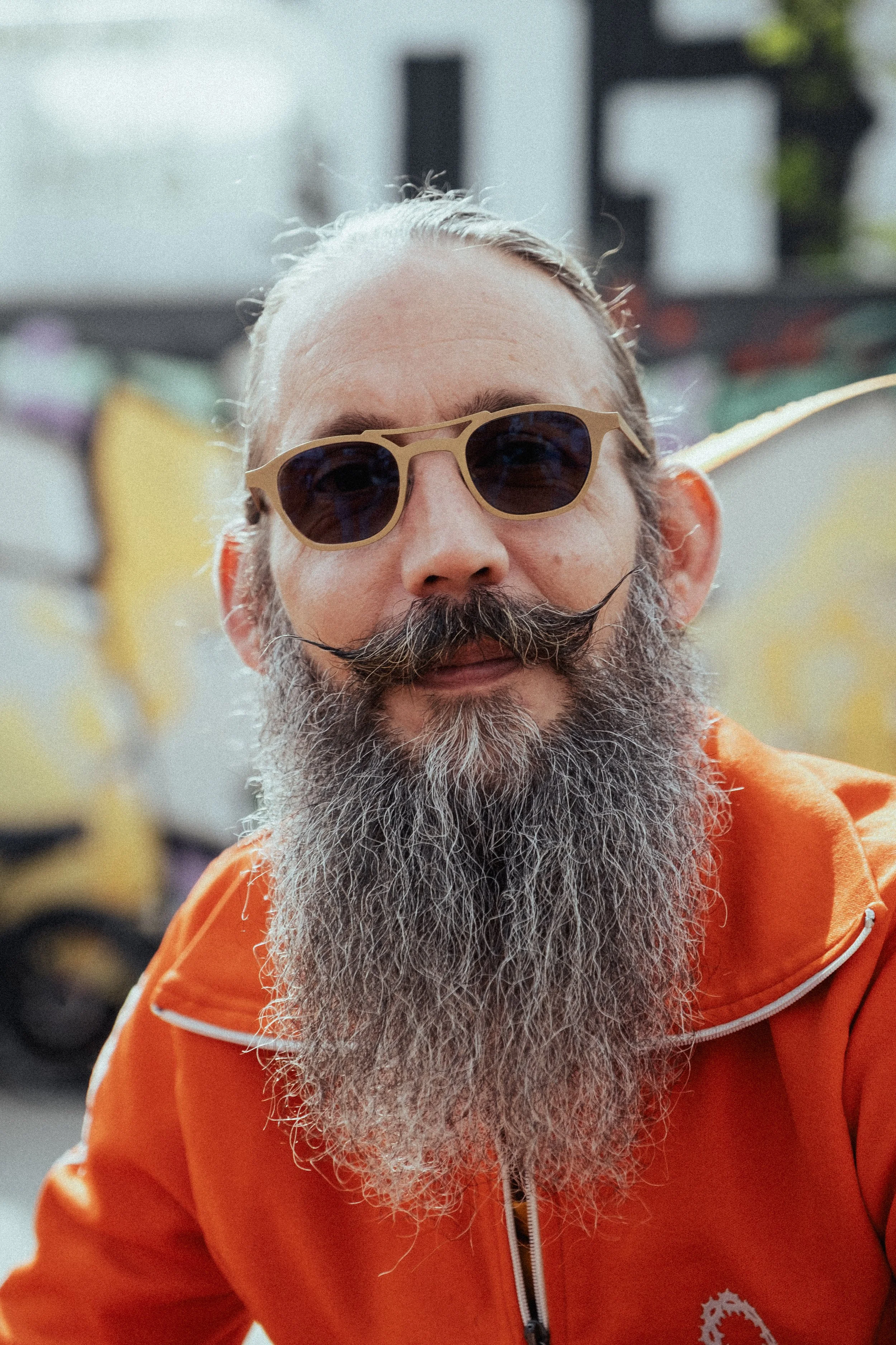 Een man met een lange, grijze baard en een snor, draagt een beige zonnebril en een oranje jack met een witte ritssluiting en een logo op de borst. De achtergrond is vaag en bevat grafitti op een muur.
