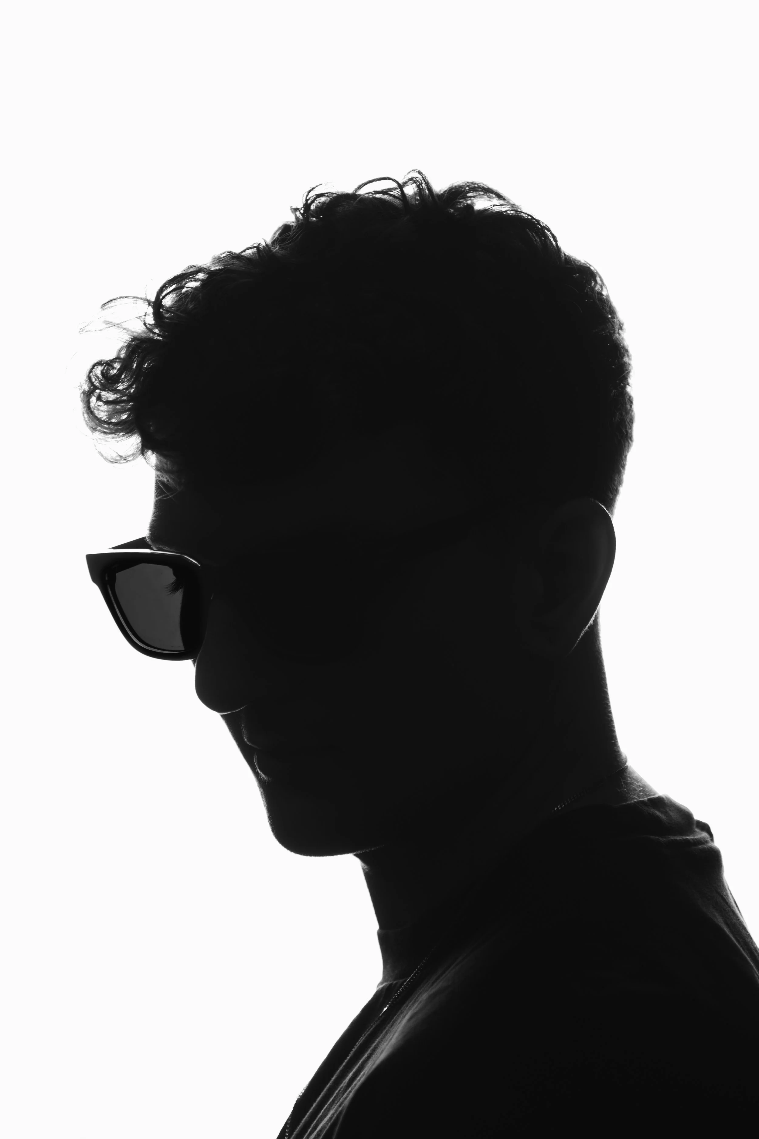 Zwart-wit silhouet van persoon met kroes haar en een zonnebril, kijkt naar rechts, witte achtergrond.