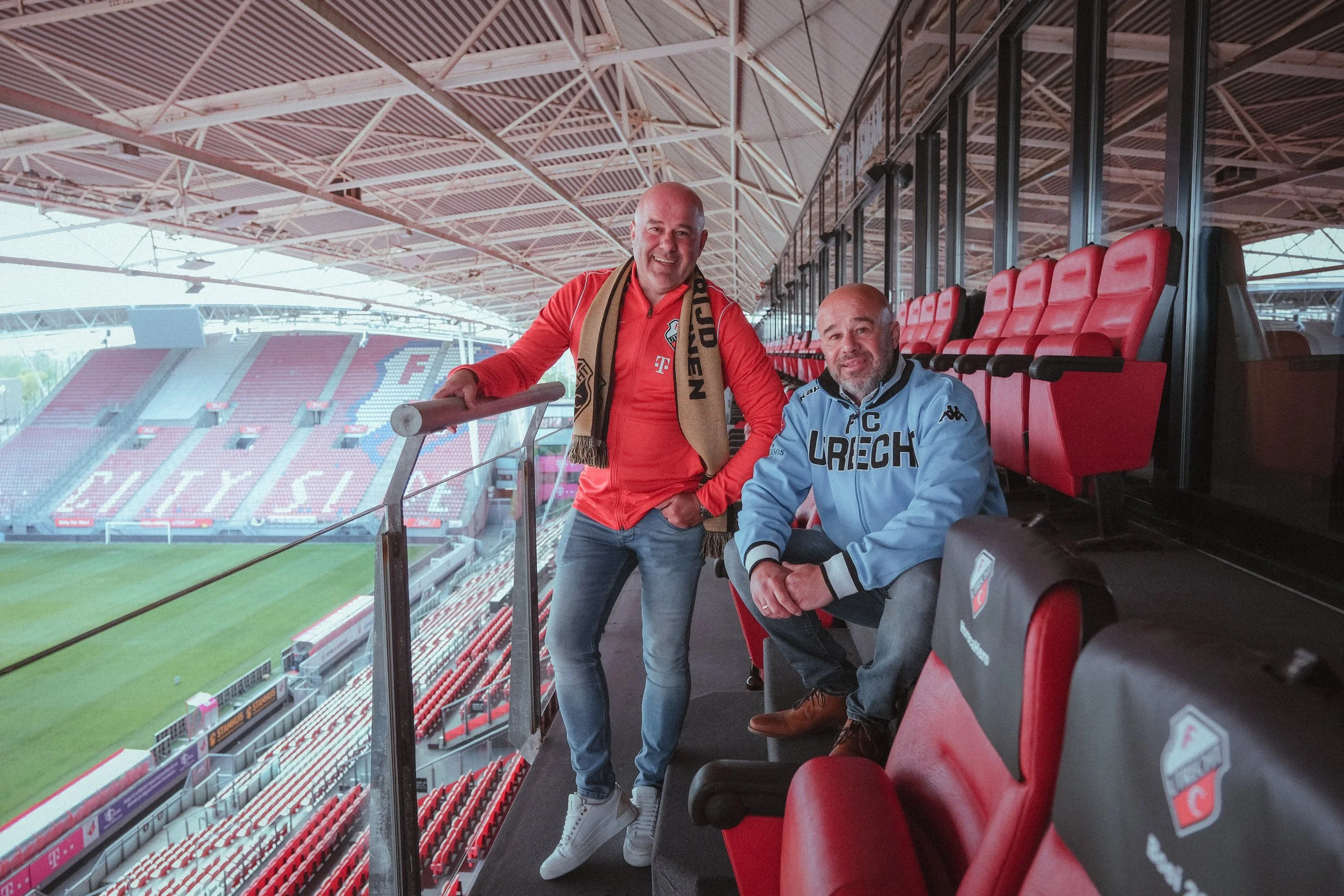 Twee mannen in een voetbalstadion, één staat en één zit, beide dragen sportkleding en lachen, met lege rode stadionstoelen op de achtergrond.