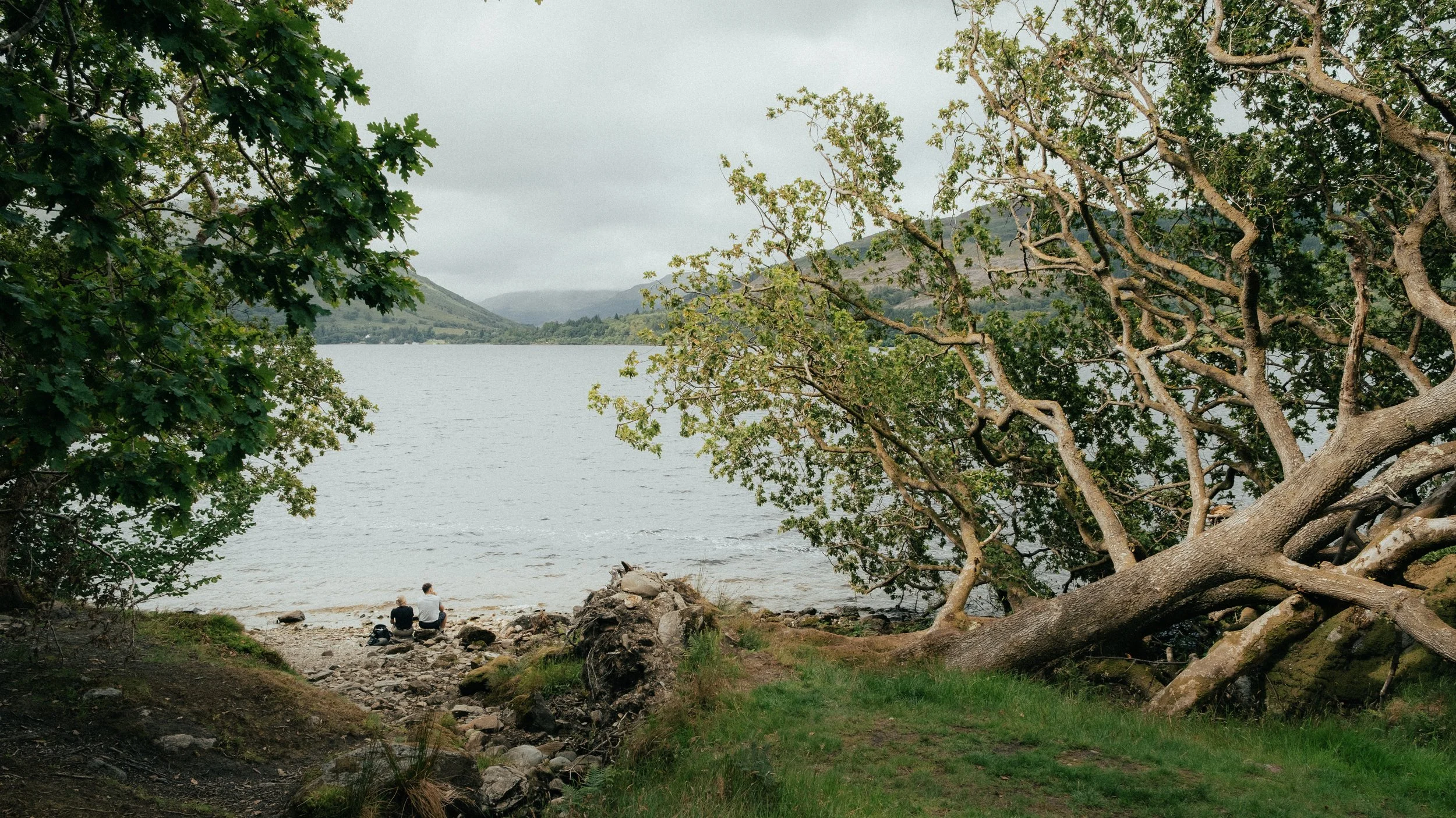 Twee mensen zittend aan de oever van een meer, omgeven door groene bomen en bergen op de achtergrond, op een bewolkte dag.