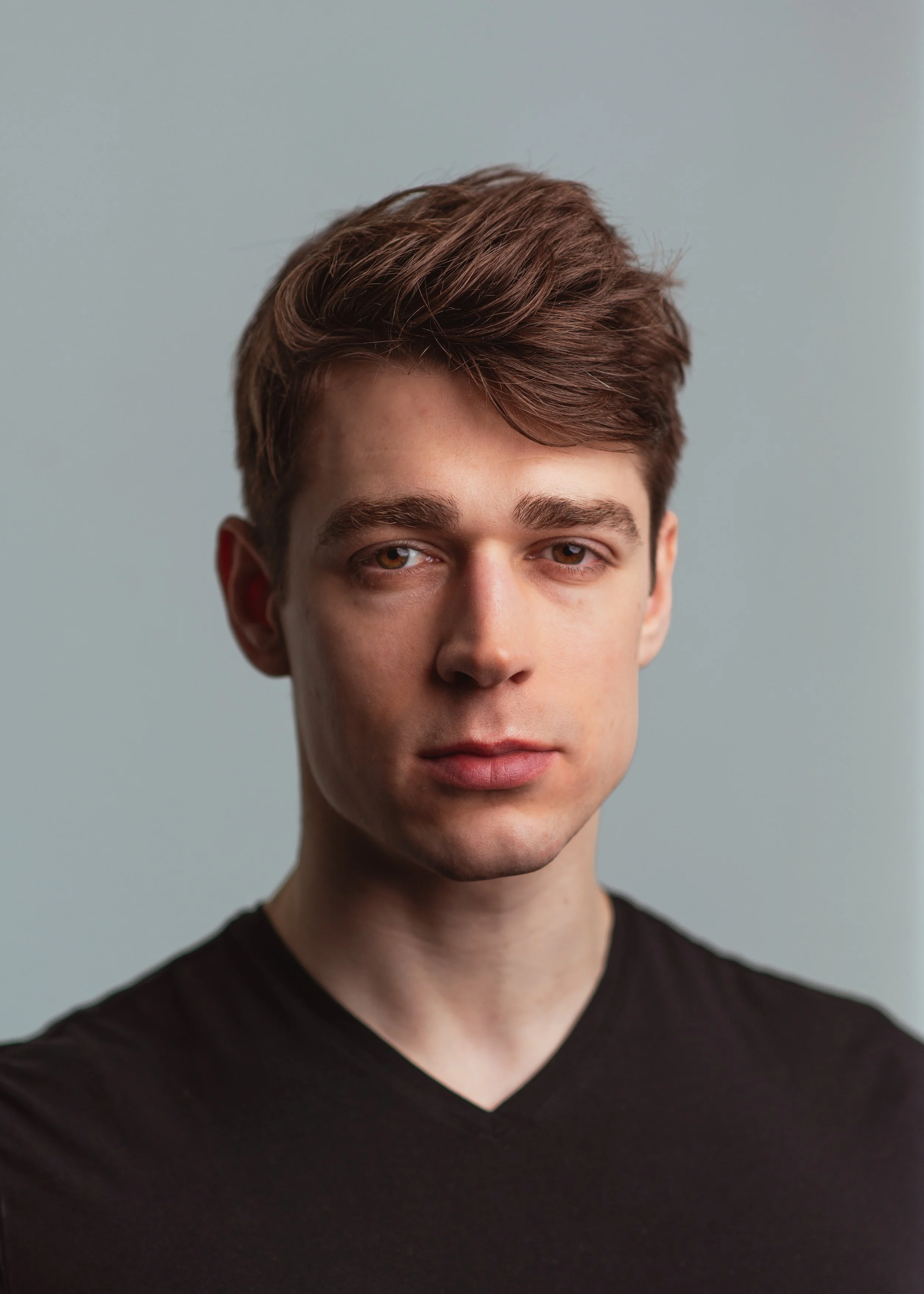 Close-up van een jonge man met bruine, golvende haar en licht getinte huid, gekleed in een zwart V-hals shirt, tegen een effen grijze achtergrond.