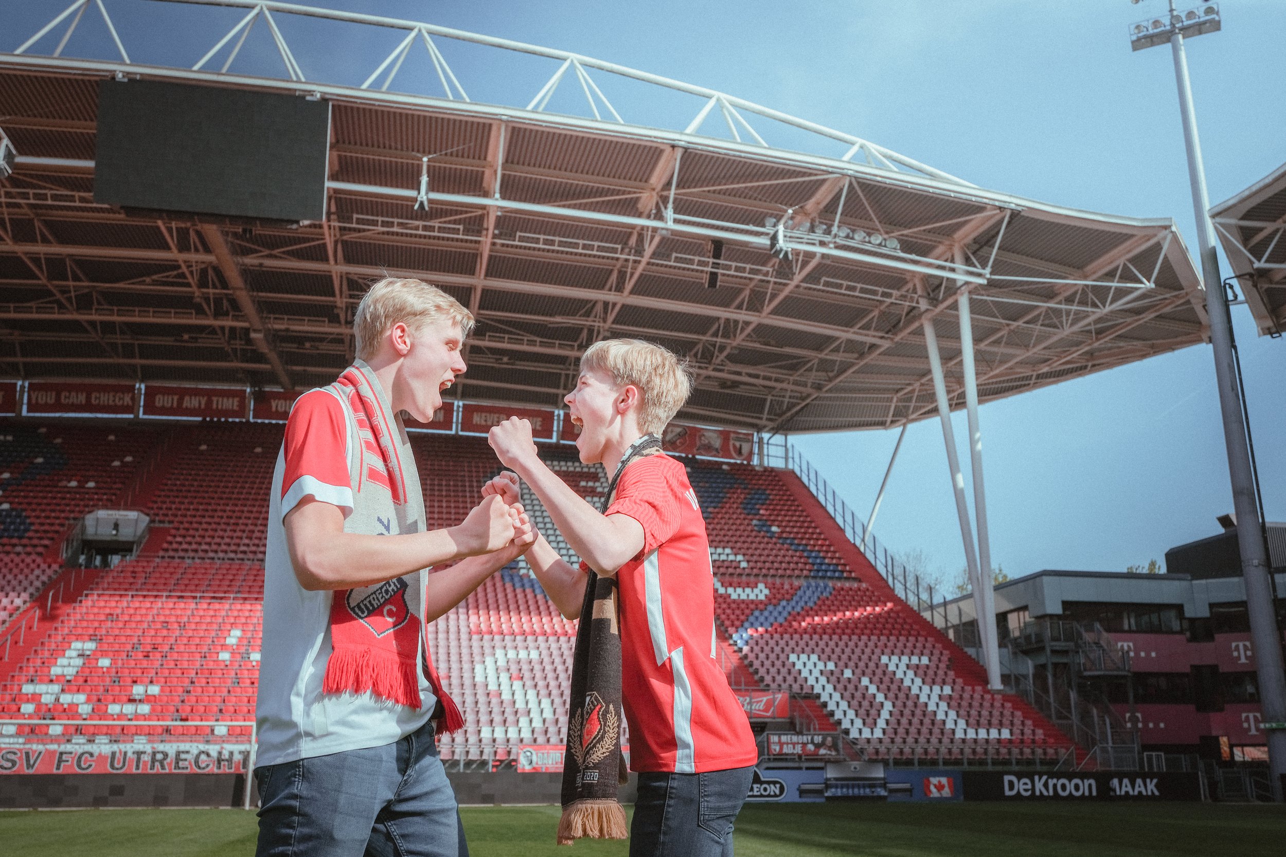 Twee jonge mannen staan op een voetbalstadion veld en schudden handen. Een van hen heeft een zwart en goud gedrapeerd sjaal om de nek. Ze lijken blij en enthousiast.
