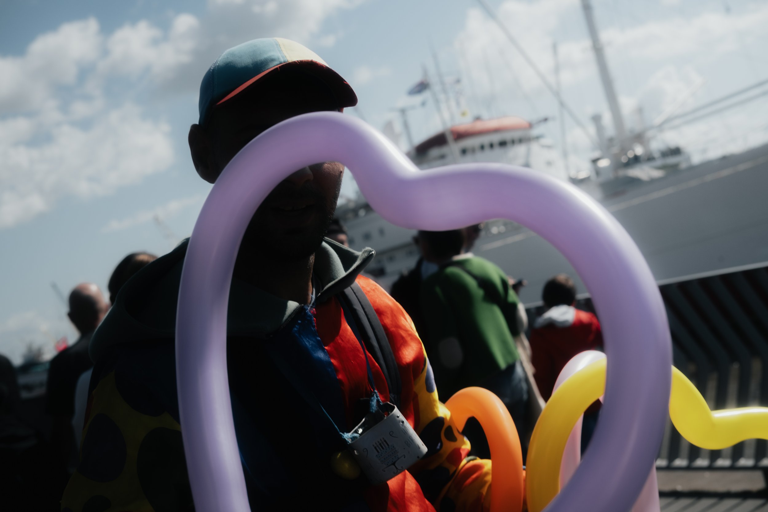 Mensen bij een evenement met ballonnen en schepen op de achtergrond onder een bewolkte hemel.