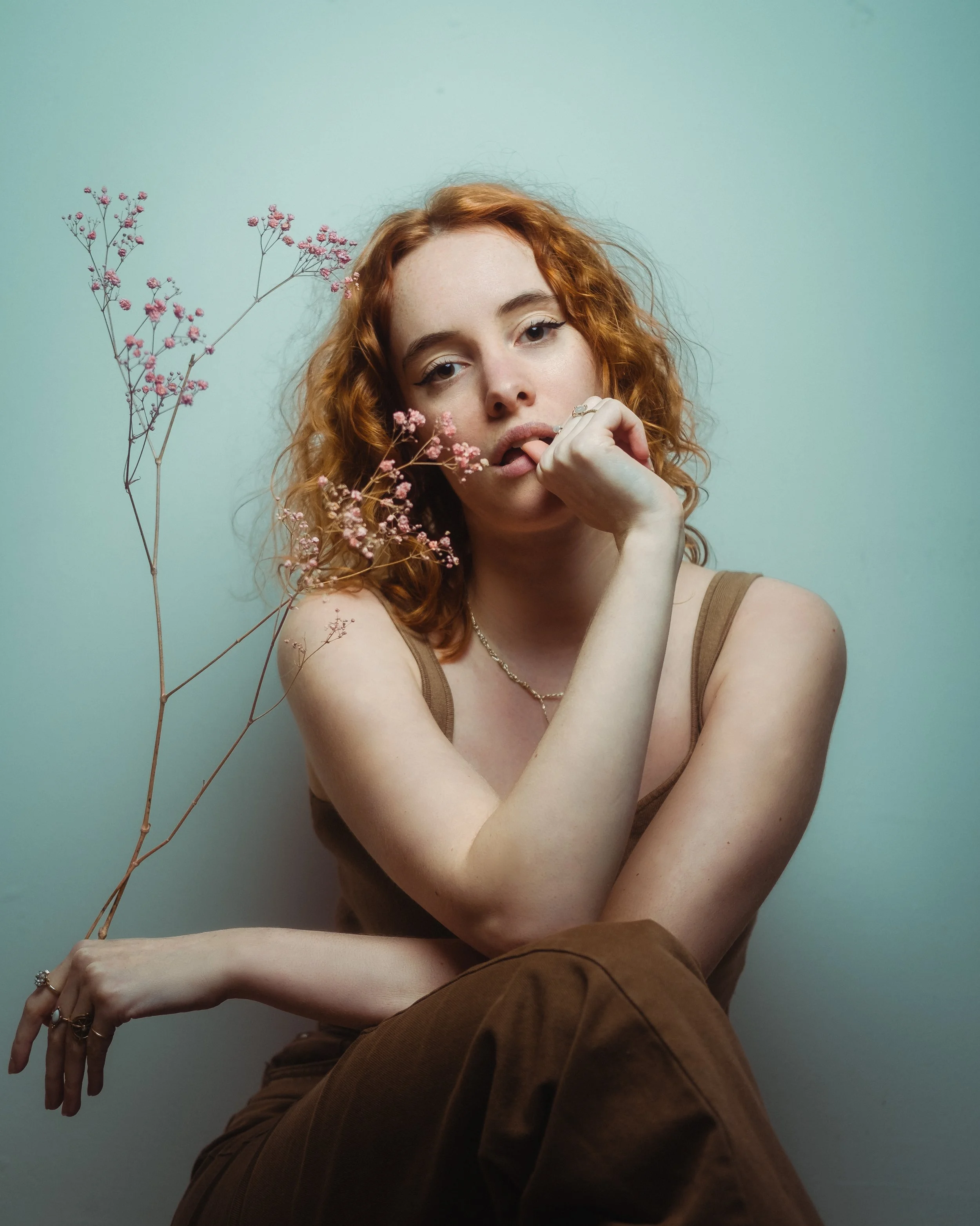 Portret van een jonge vrouw met rood, krullend haar en lichte make-up, inclusief eyeliner, met een neiging tot zijwaartse blik, die een bloemstuk met roze bloemen vasthoudt. Ze draagt een bruin topje en sieraden en zit tegen een lichtgroene achtergro