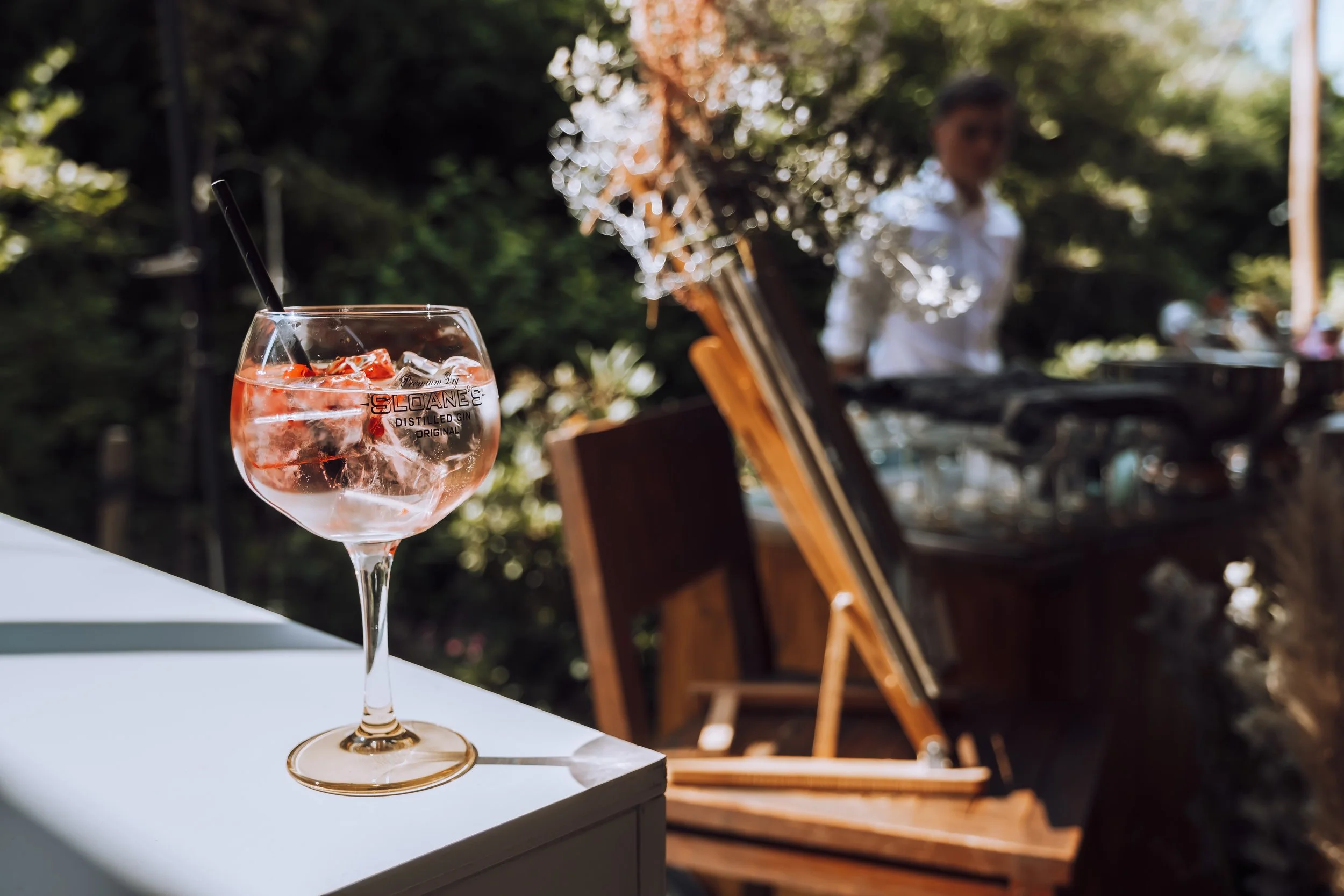 Een glas met een roze drankje, ijsblokjes en een rietje, geplaatst op een witte tafel. Op de achtergrond is een man te zien die achter een muziekinstrument speelt, met groene bomen en een blauwe lucht omheen.