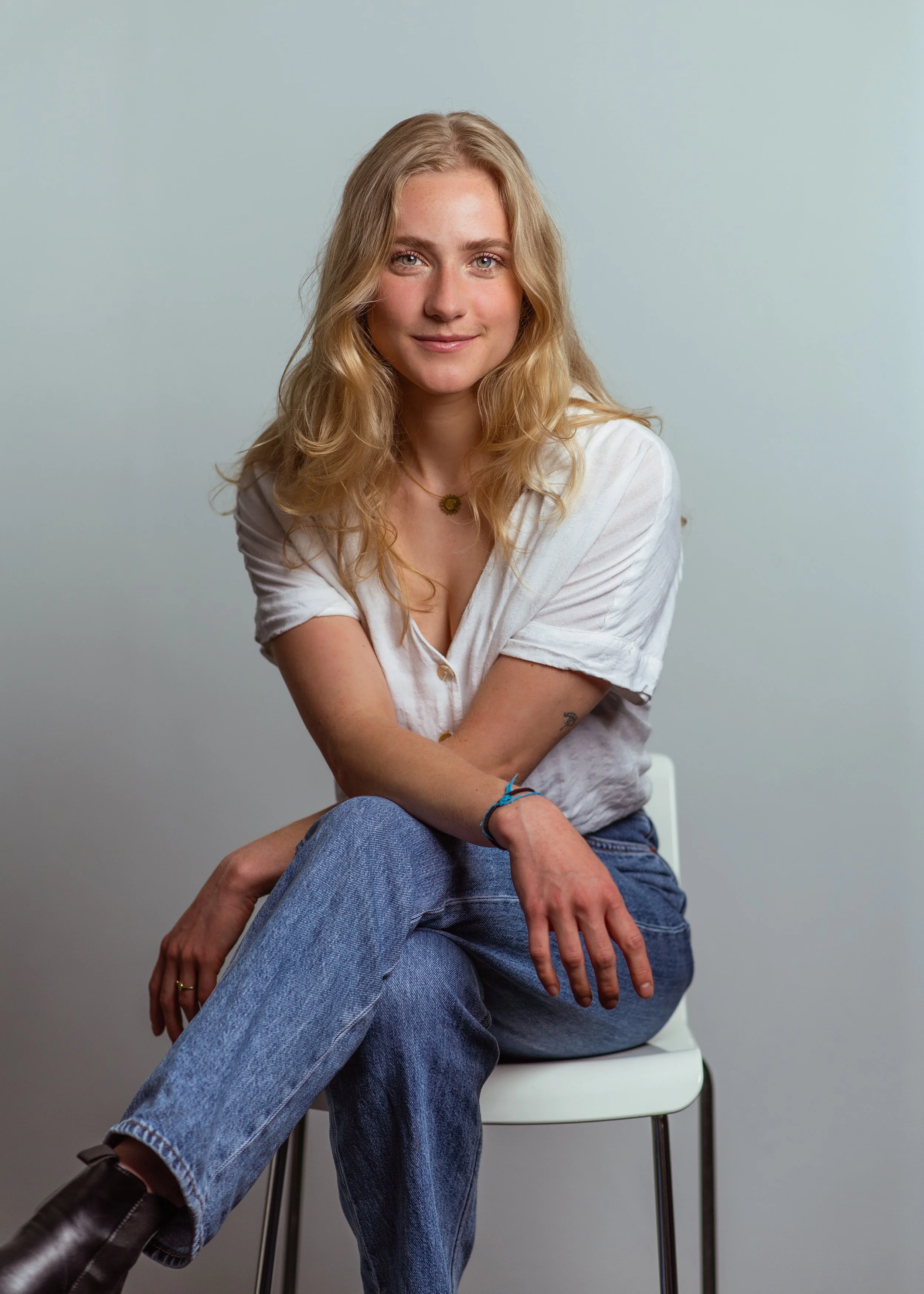 Een jonge vrouw met geblondeerd haar en blauwe ogen zit op een witte stoel tegen een lichtblauwe muur, draagt een witte blouse en blauwe spijkerbroek, en kijkt vriendelijk in de camera.