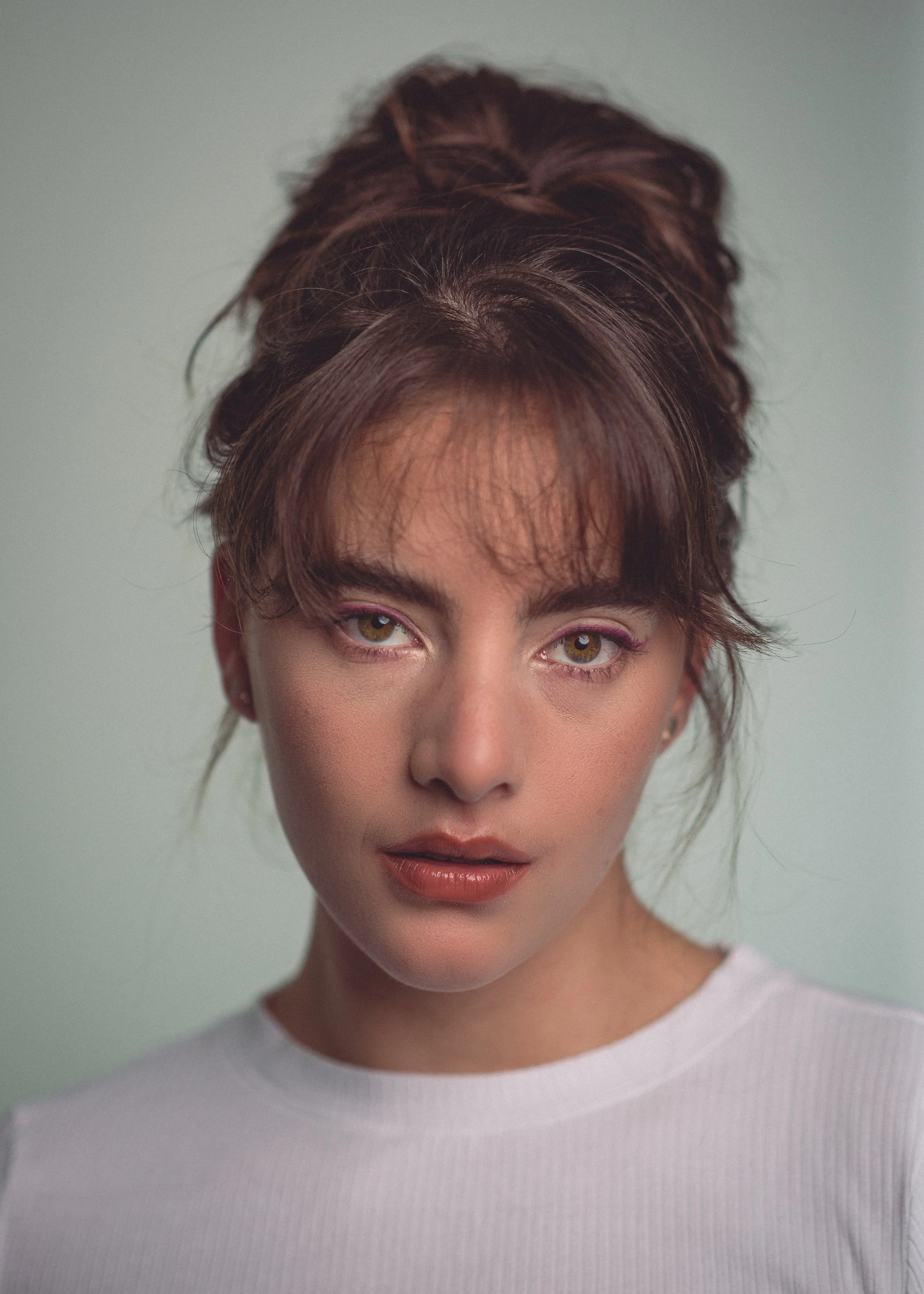 Portret van een vrouw met bruin haar en groene ogen, gekleed in een witte top, met een neutrale uitdrukking en een zachte schemerlichte achtergrond.