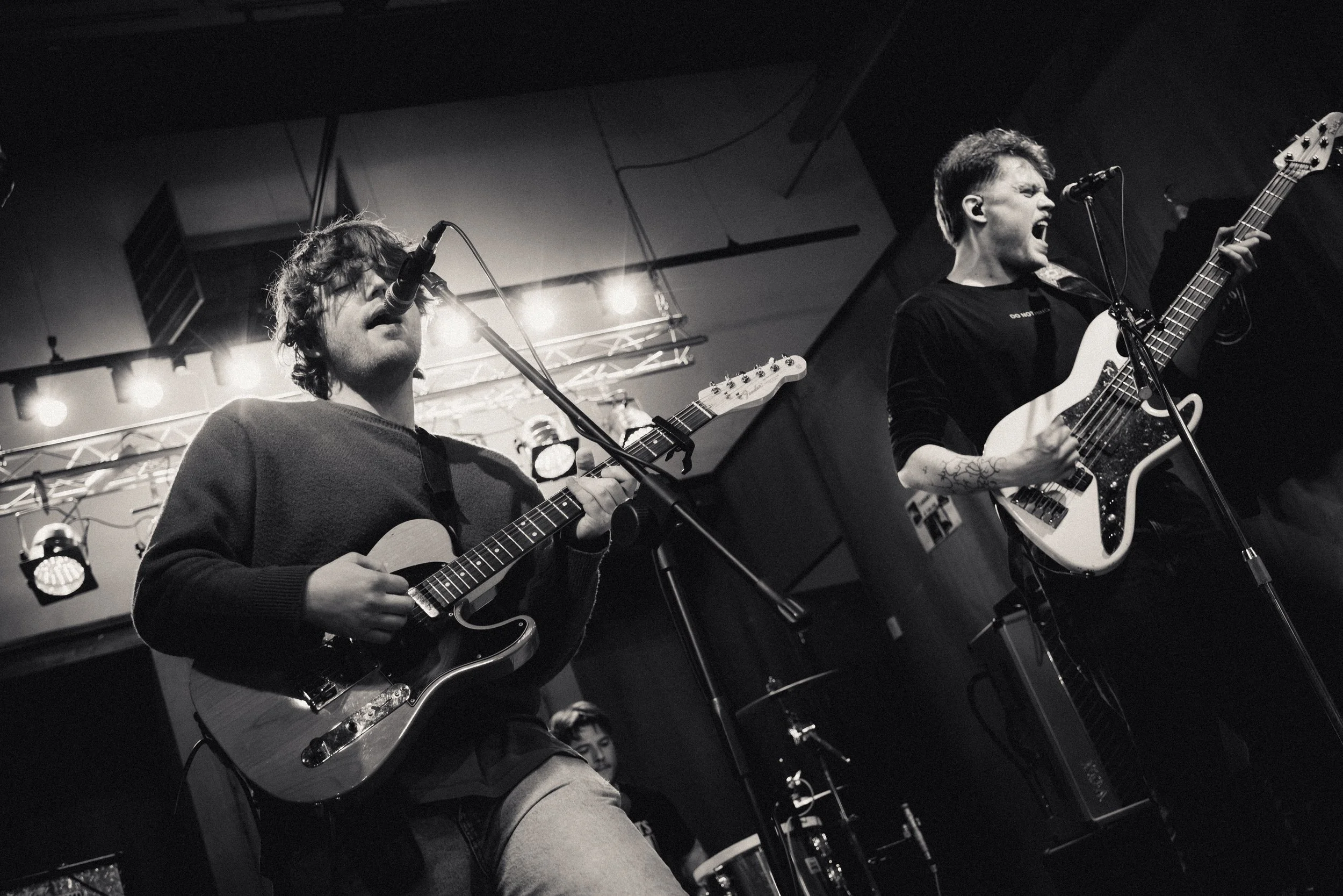 Twee muzikanten spelen gitaar en basgitaar tijdens een live optreden in een donkere zaal, zwart-wit foto.