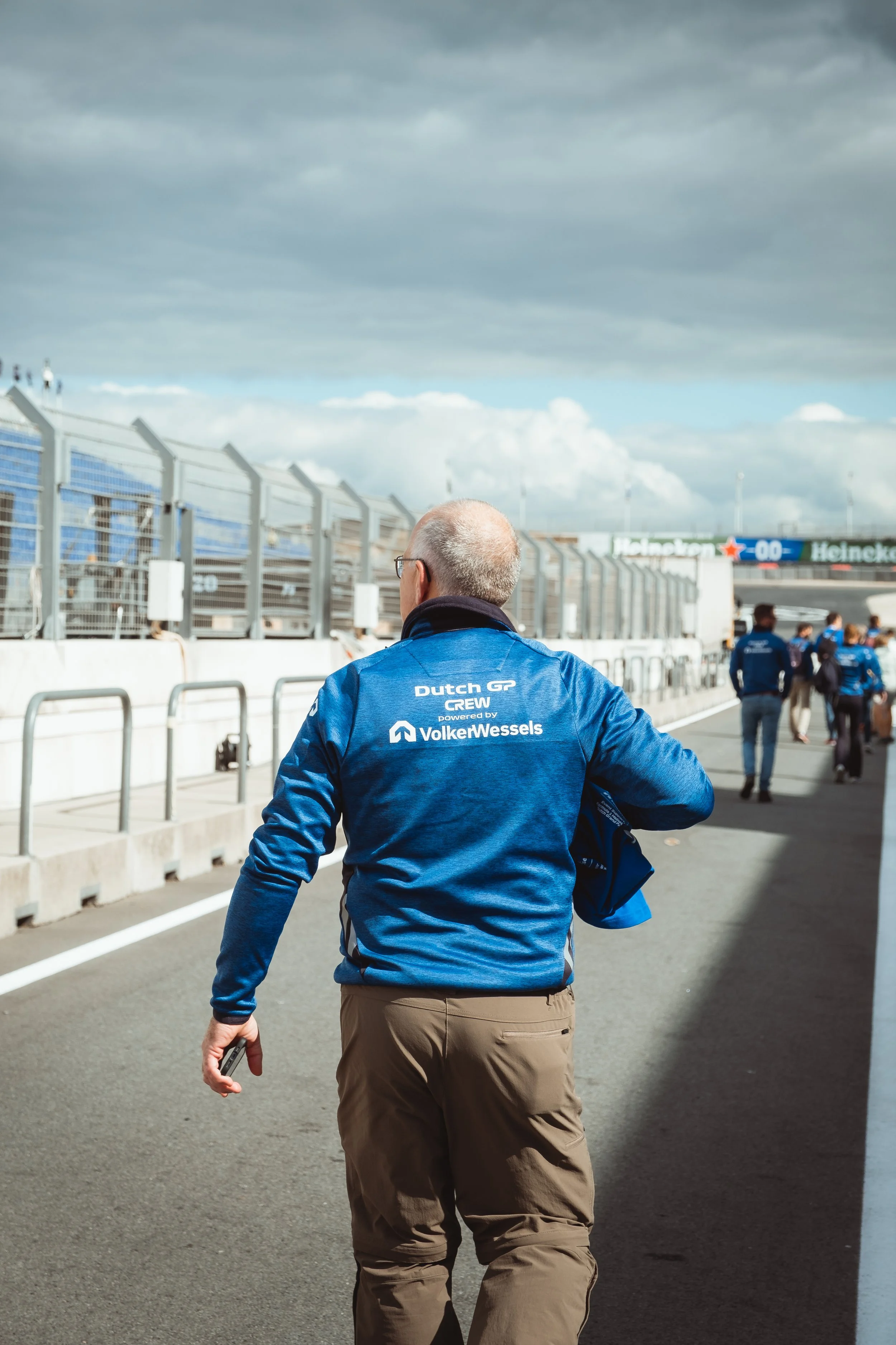 Man in blauwe jas met tekst 'Dutch GP CREW powered by VolkerWessels' wandelt op een circuit, met andere mensen op de achtergrond onder donkere wolken.