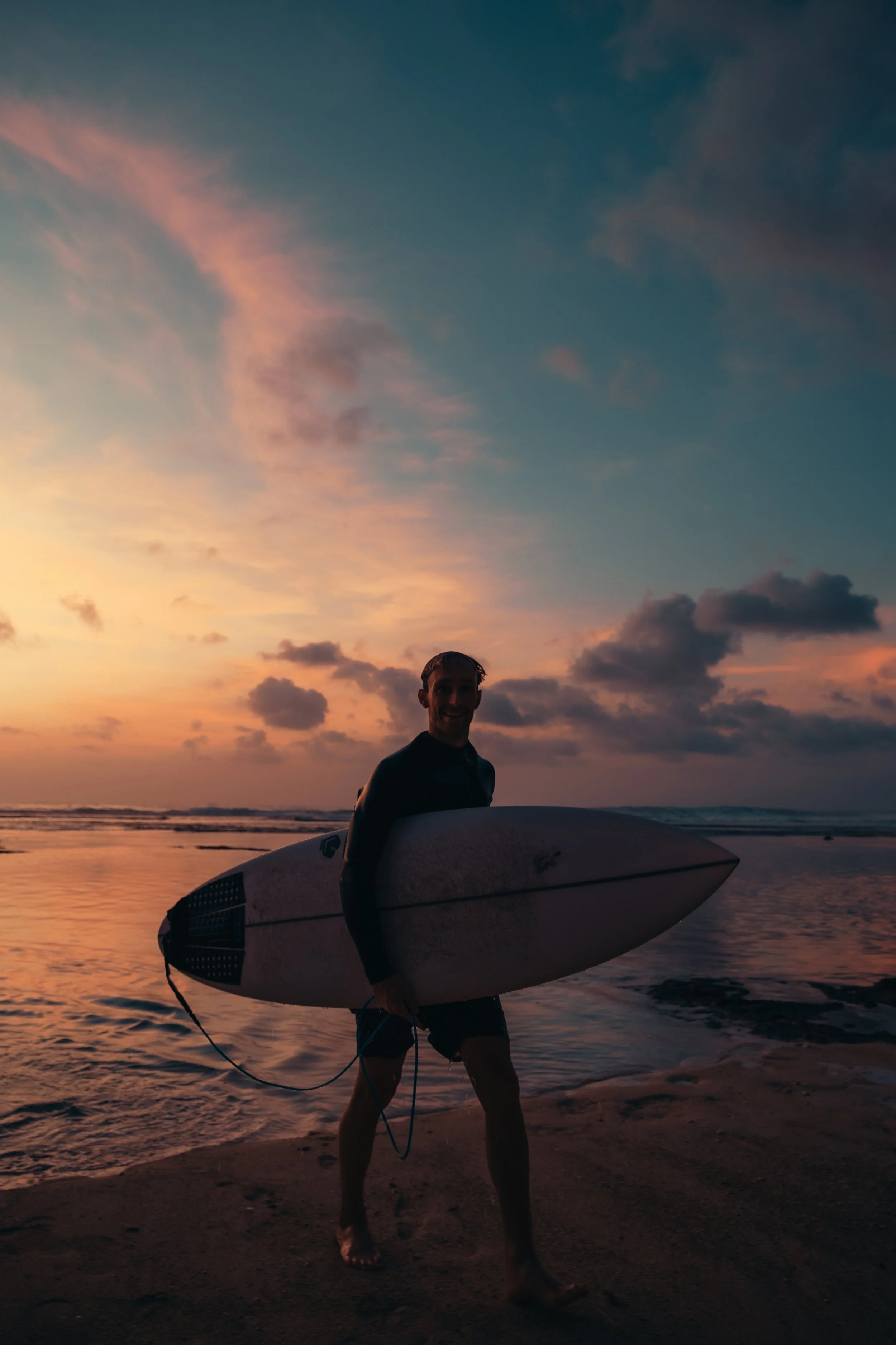 Een man met surfboard loopt op het strand bij zonsondergang, de lucht is roze en blauw, de zee golft zachtjes.