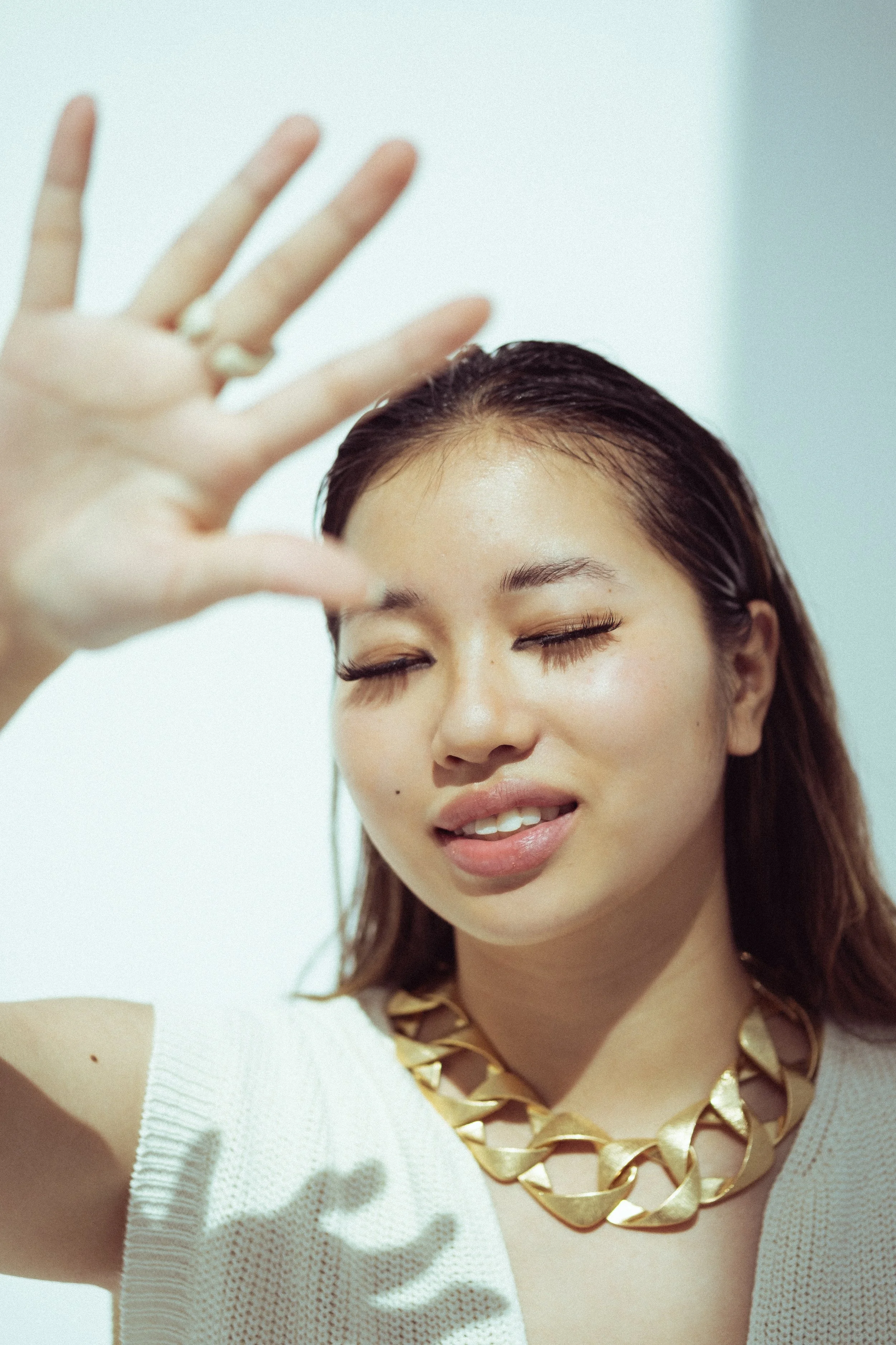 Close-up van een glimlachende vrouw met bruin haar, oorbelletjes, en een grote gouden ketting, met haar ogen dicht en een hand omhoog.