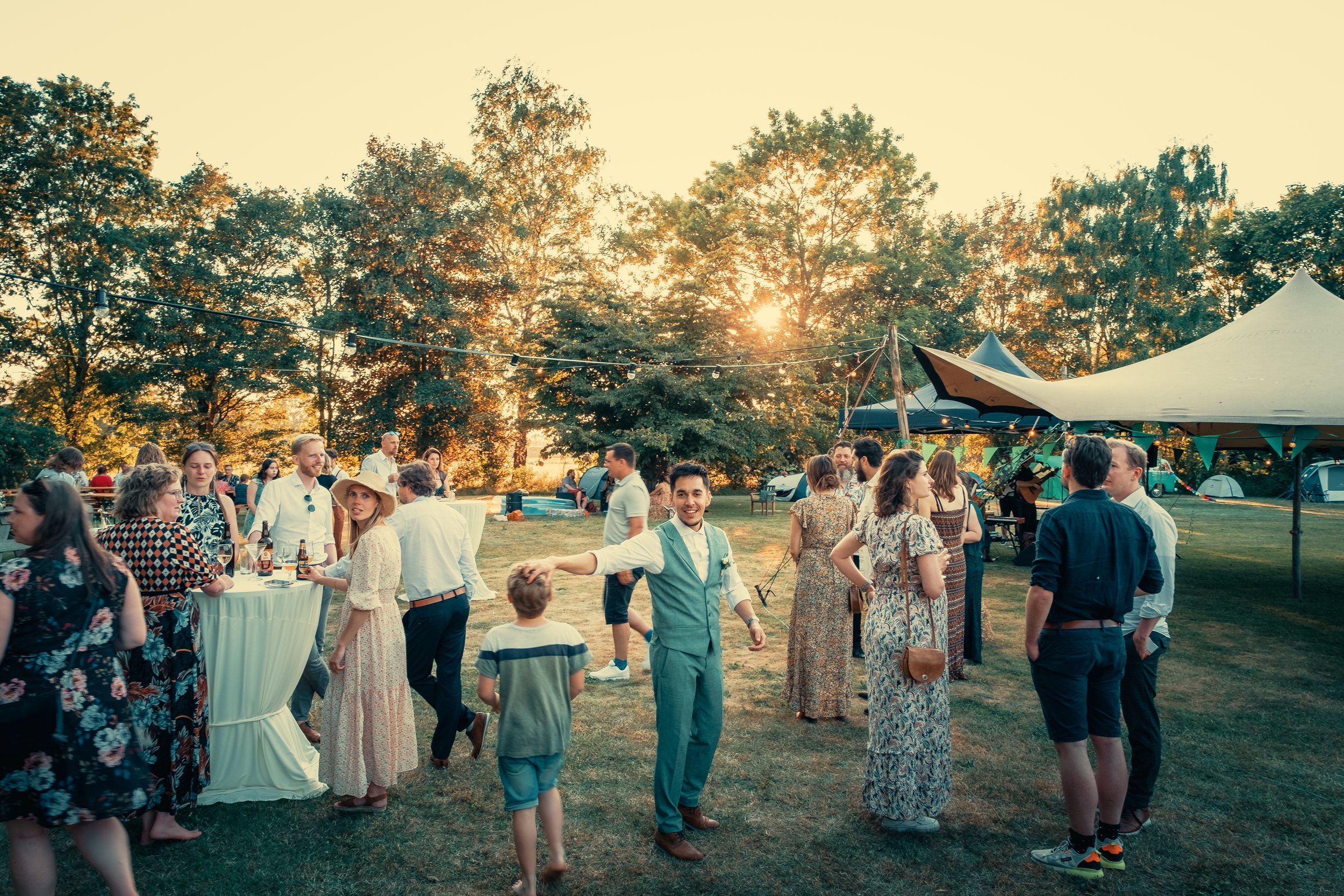 Mensen genieten van een buitenfeest in de namiddag met tafels, tenten en verlichting, rond een park met bomen tijdens zonsondergang.