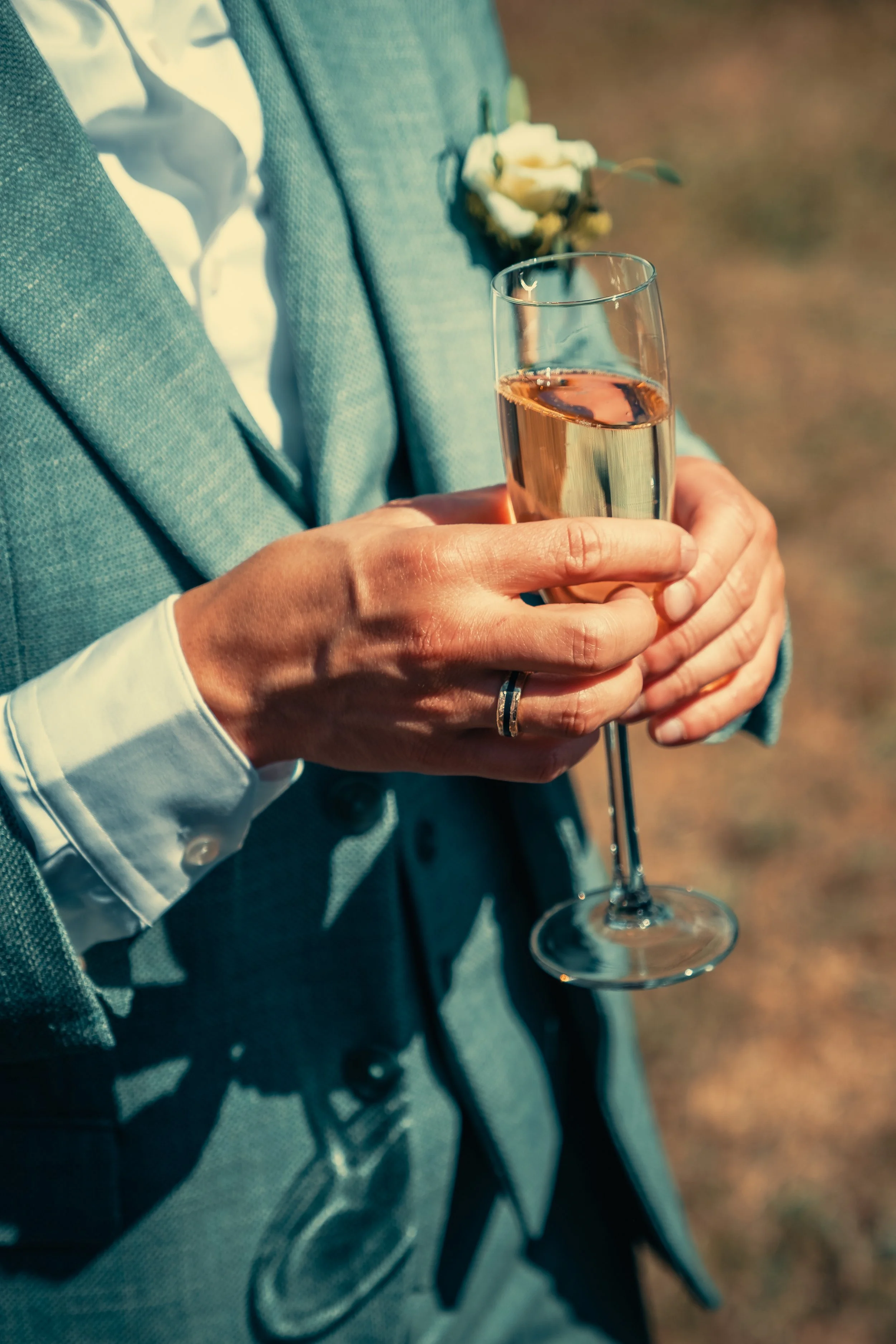 Een persoon in een grijs jasje en witte overhemd houdt een glas champagne vast, met een corsage bevestigd op de jas. De achtergrond is vaag en buiten.