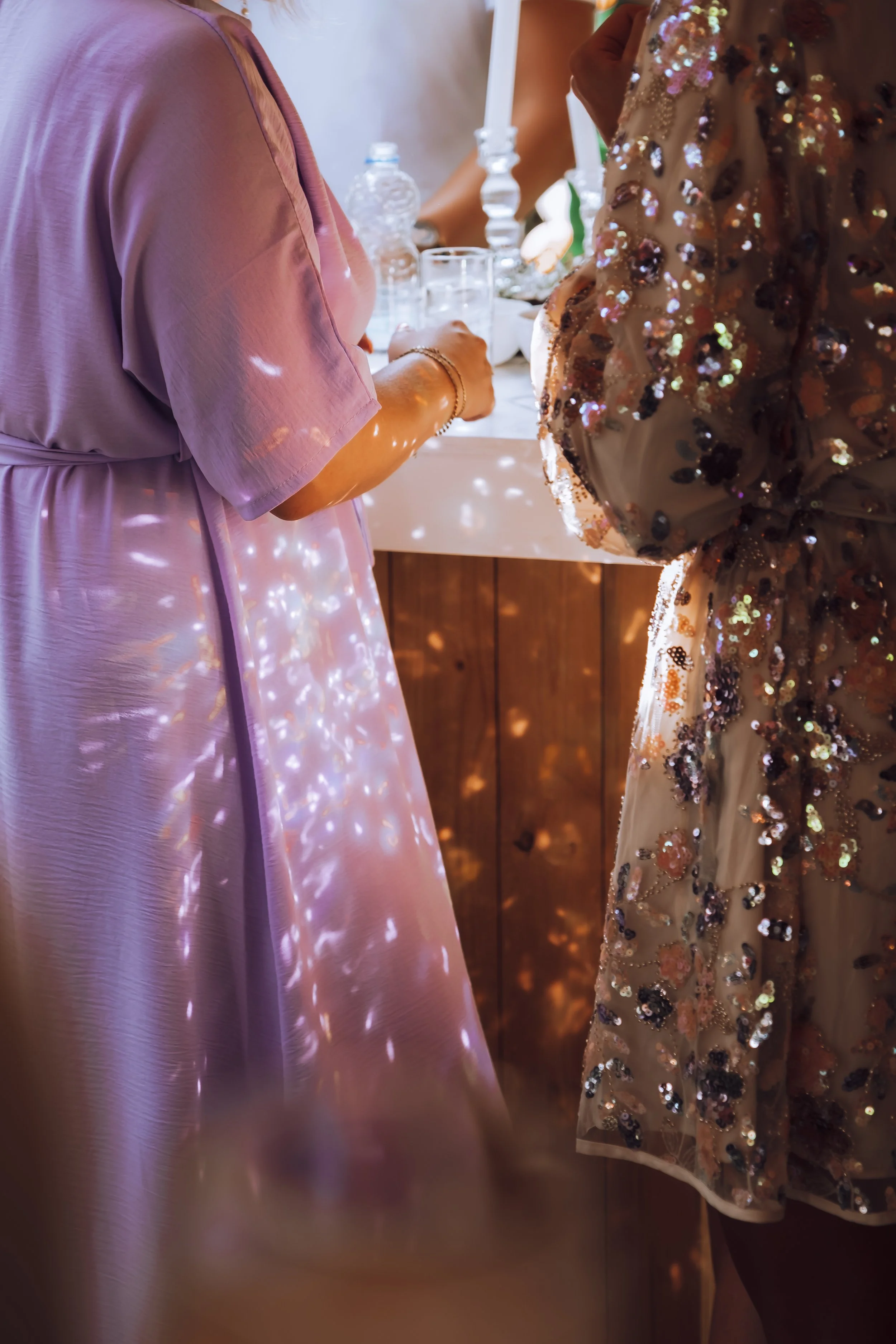 Close-up van mensen in feestkleding bij een tafel met drank en kaarsen, met glitter en lichteffecten op de kleding en omgeving