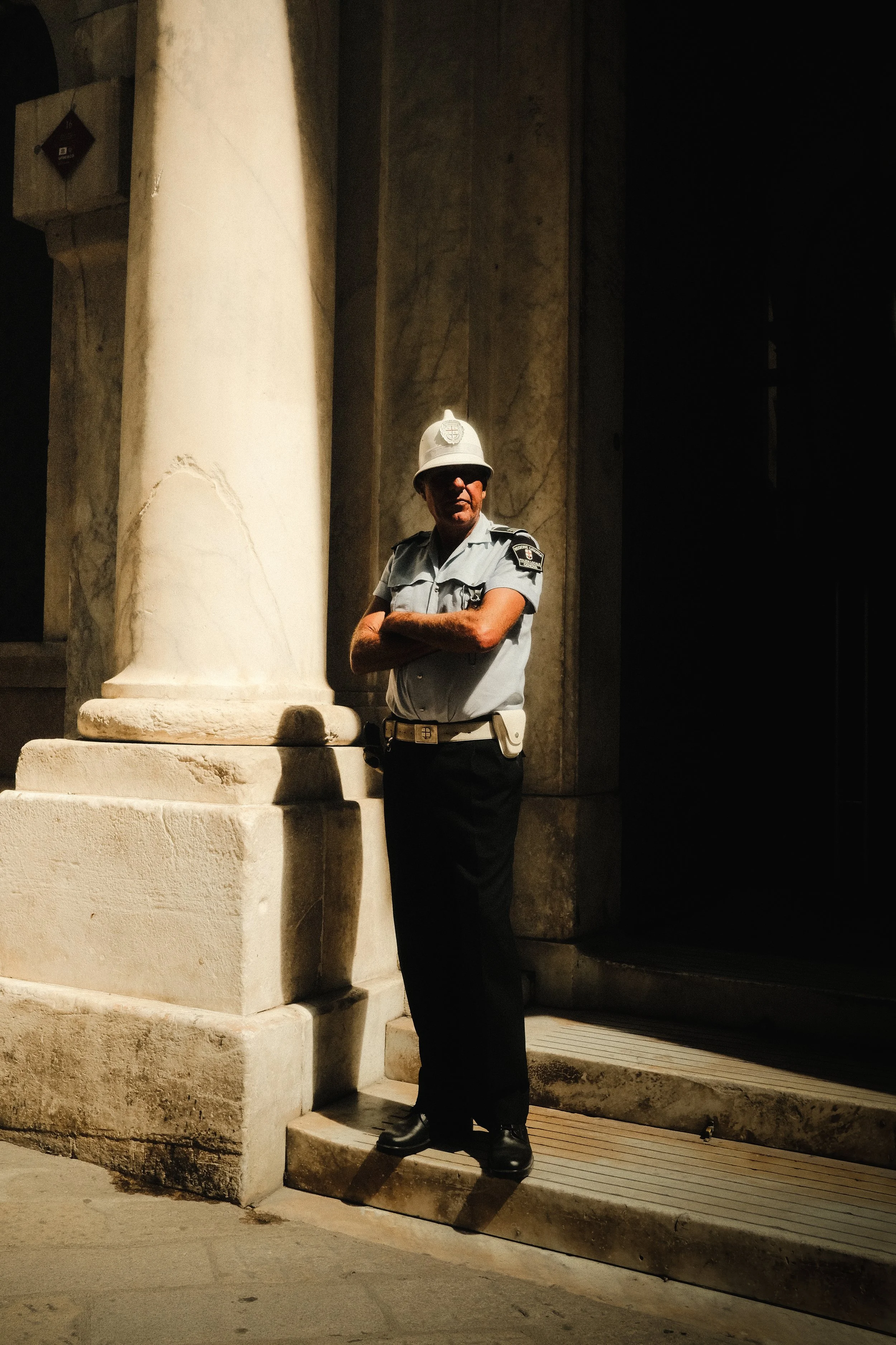 Politieagent staat met gekruiste armen naast een grote marmeren pilaar, gekleed in een lichtblauw uniform met een politiepet, donkere broek en laarzen, terwijl hij schittert in het donker.