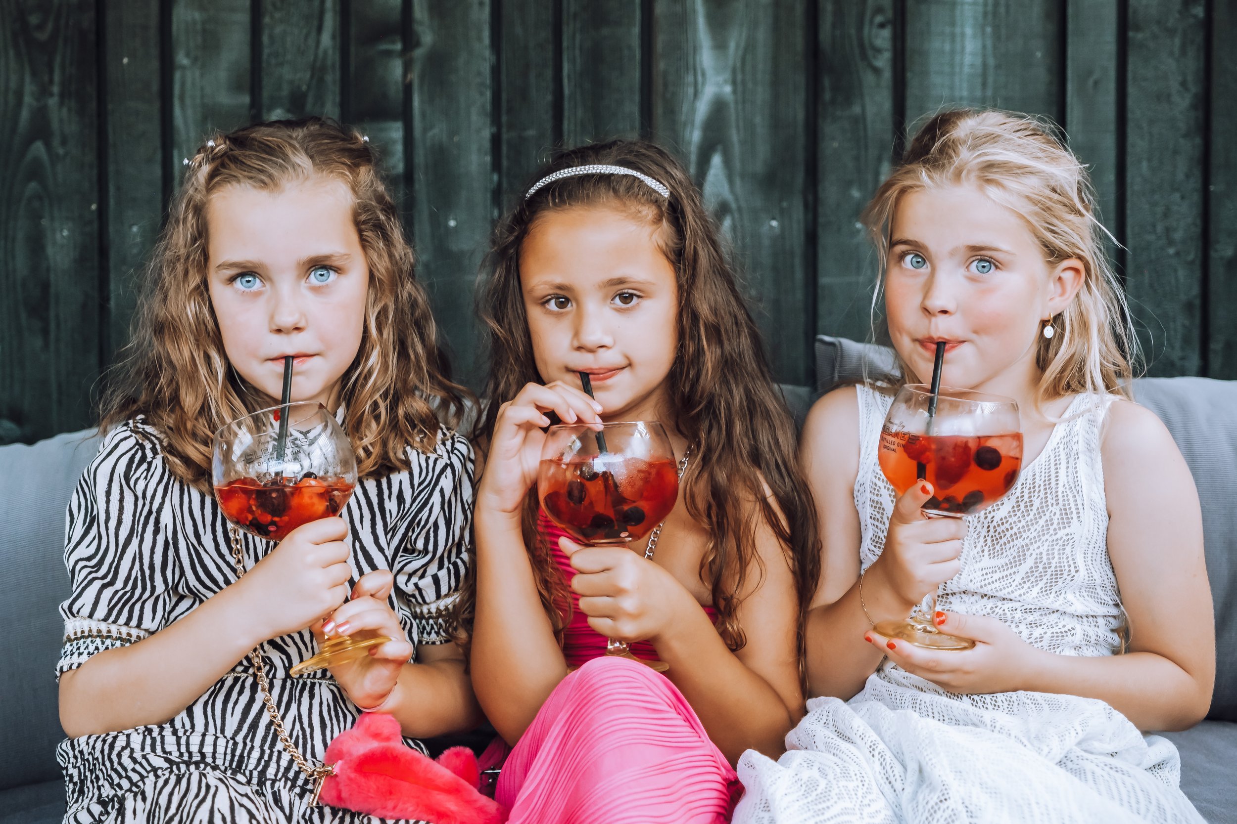 Drie jonge meisjes met blonde en bruin haar drinken cocktails met bessen en ijs, zittend op een bank tegen een houten wand.