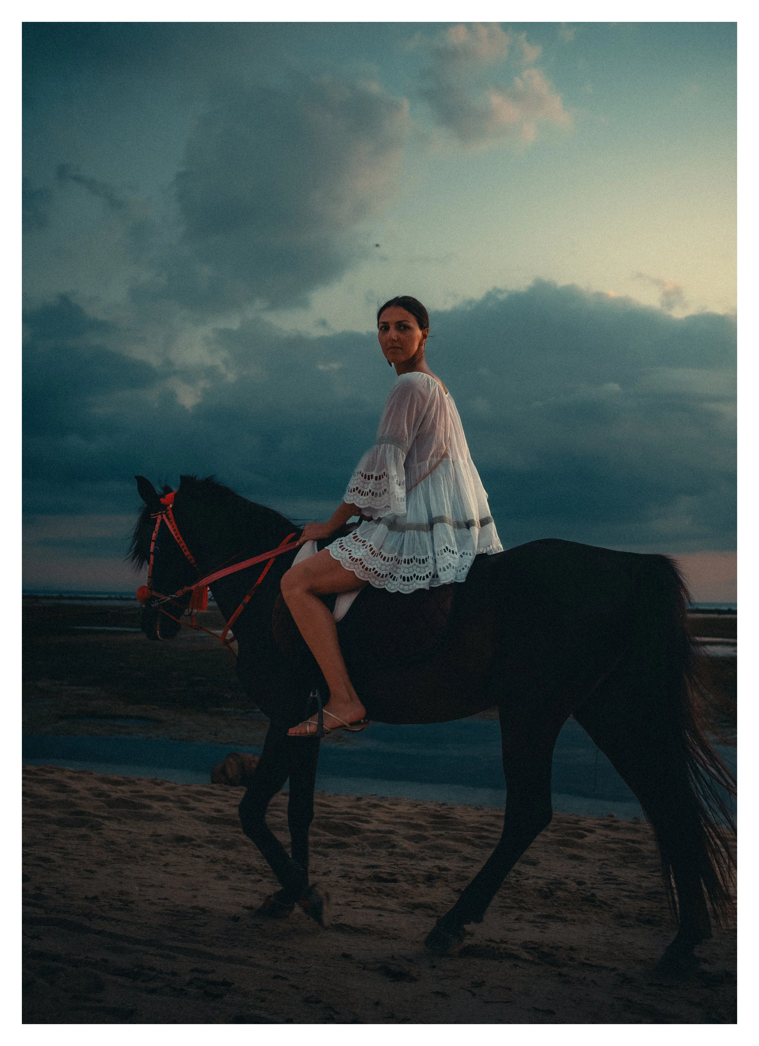 Vrouw zit te paard op het strand tijdens zonsondergang, gekleed in een witte jurk met opengewerkte details, met een bewolkte hemel op de achtergrond.