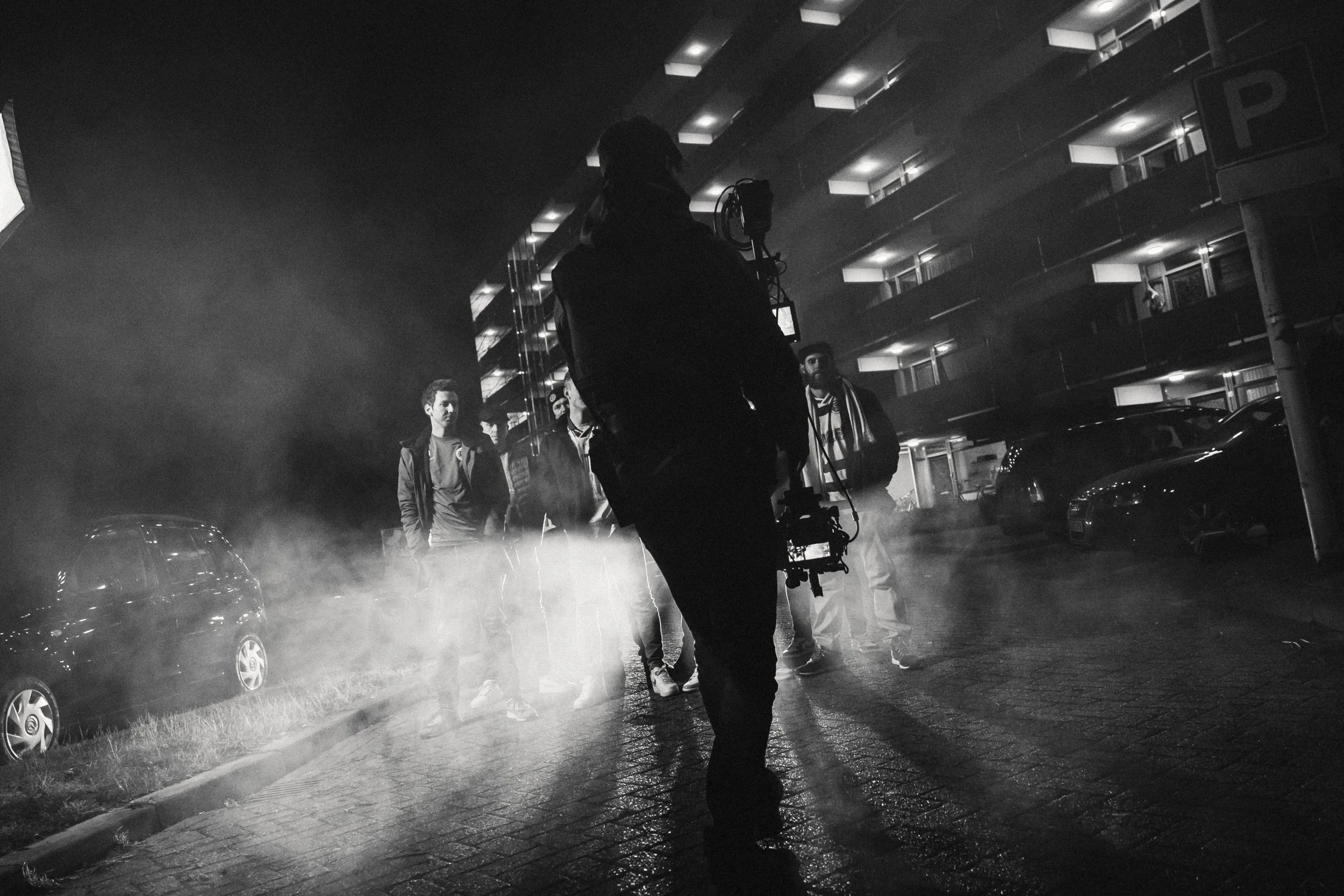 Zwart-wit foto van een groep mensen die op straat staan bij nacht, met een persoon in de voorgrond die een camera of filmingapparaat vasthoudt, en een appartementencomplex op de achtergrond.