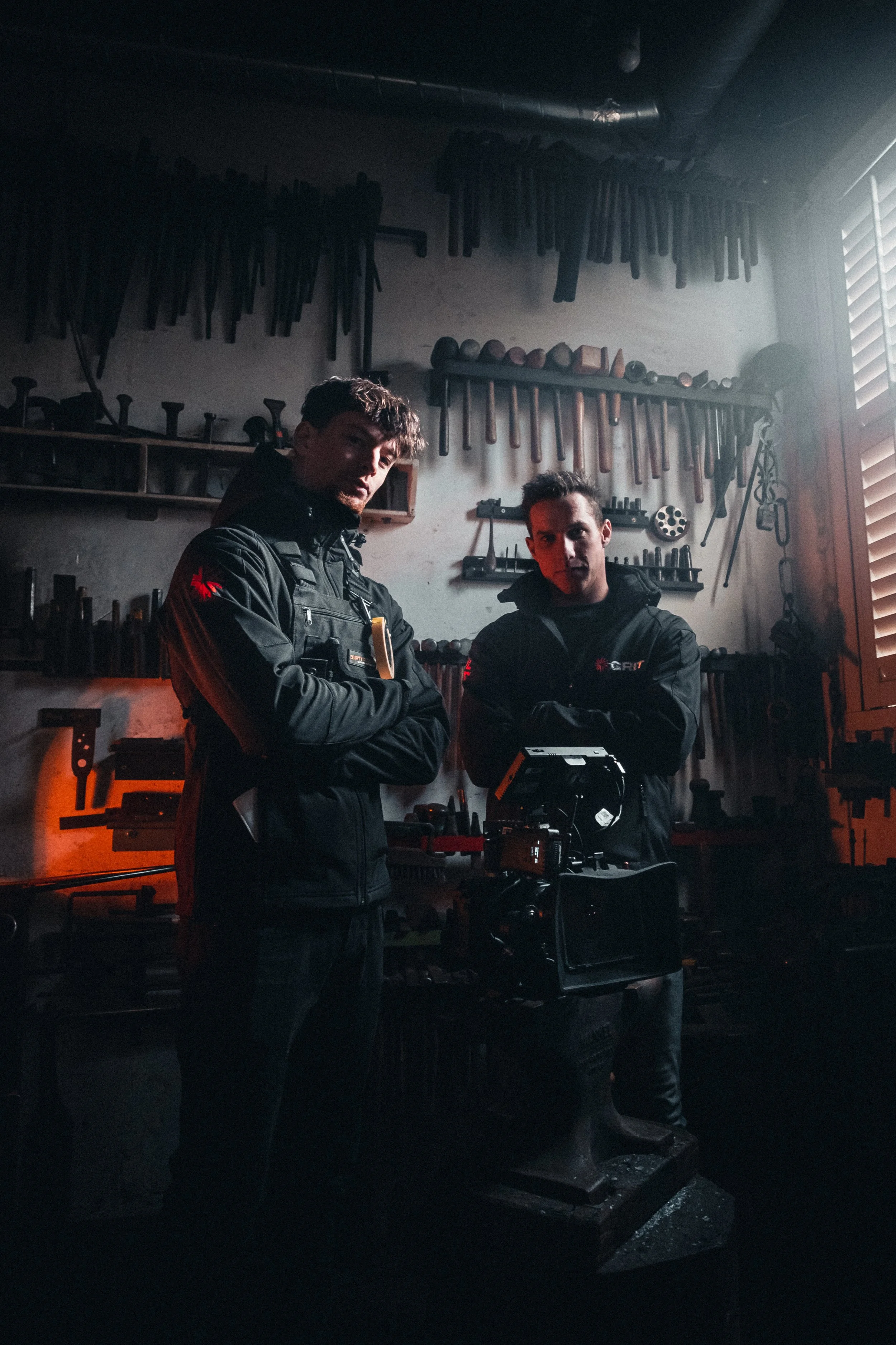 Twee mannen in een werkplaats met gereedschap aan de muur, één van hen houdt een filmcamera vast.