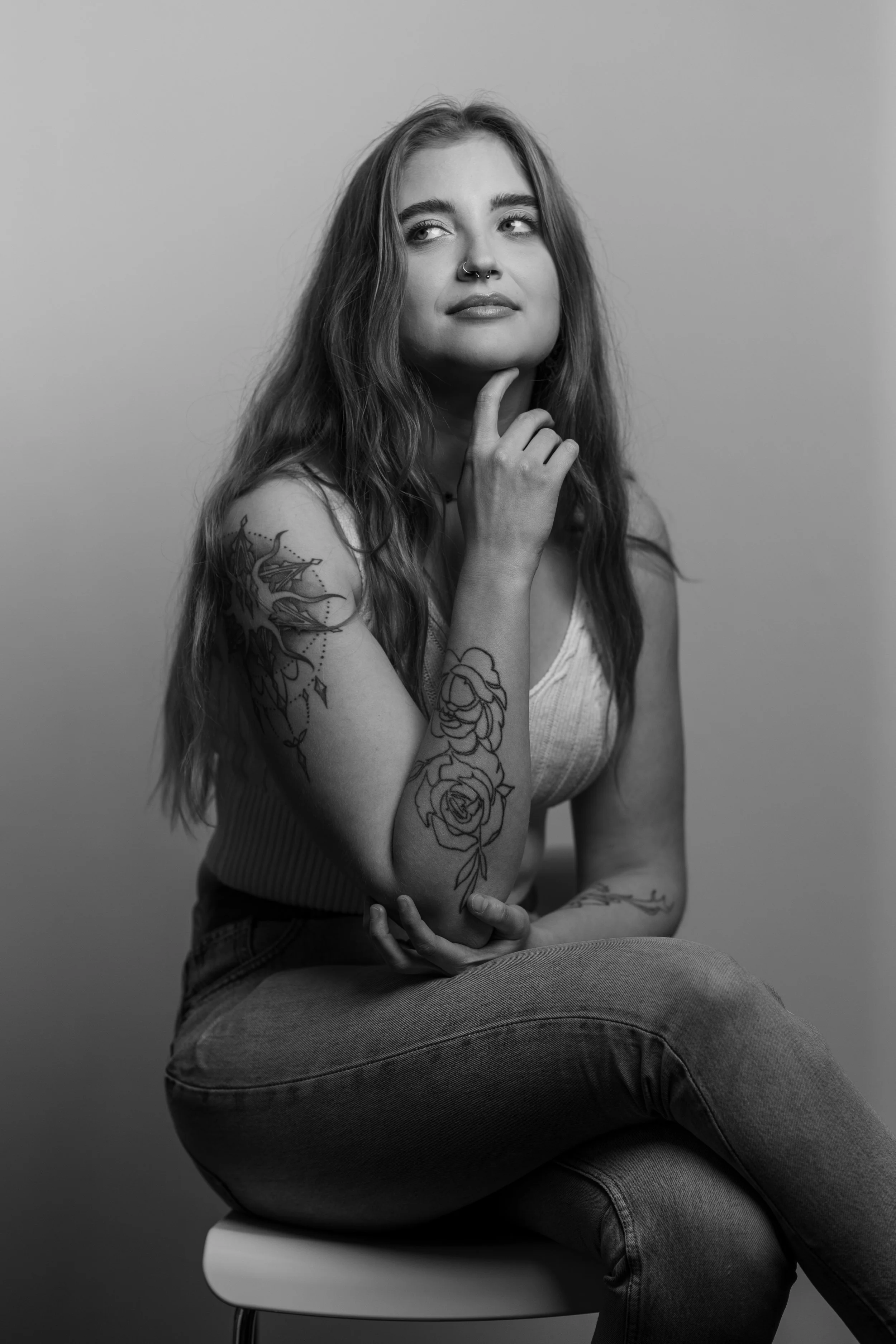 Zwart-wit foto van een jonge vrouw met tatuaties op haar arm, zittend op een witte stoel, met haar kin op haar hand, die over haar borst ligt, tegen een effen achtergrond.