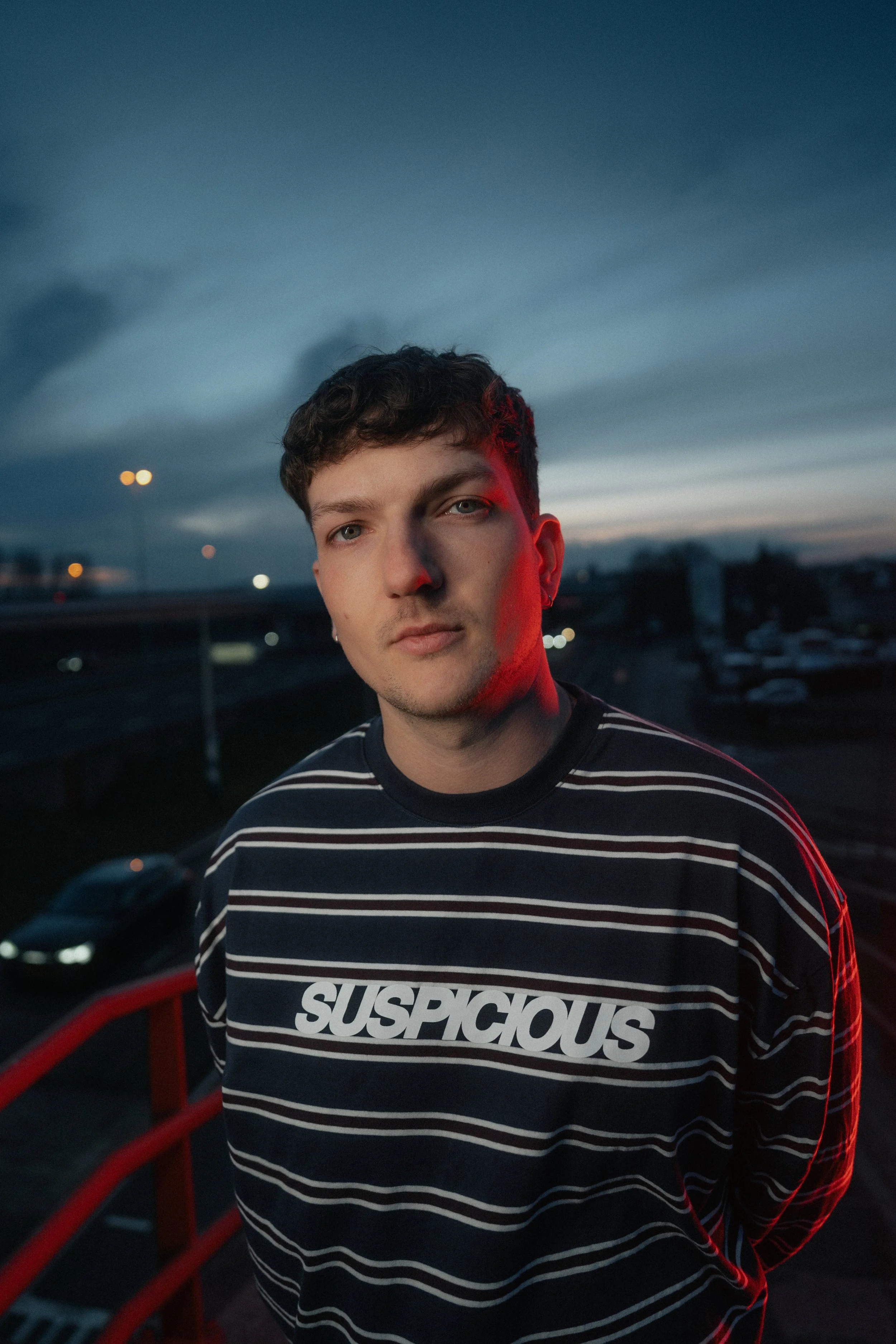 Portret van een jongeman met kort, gekruld haar, dragend een zwart-wit gestreept shirt met de tekst 'SUSPCIOUS' en een blauwe achtergrond met wolken bij zonsondergang.