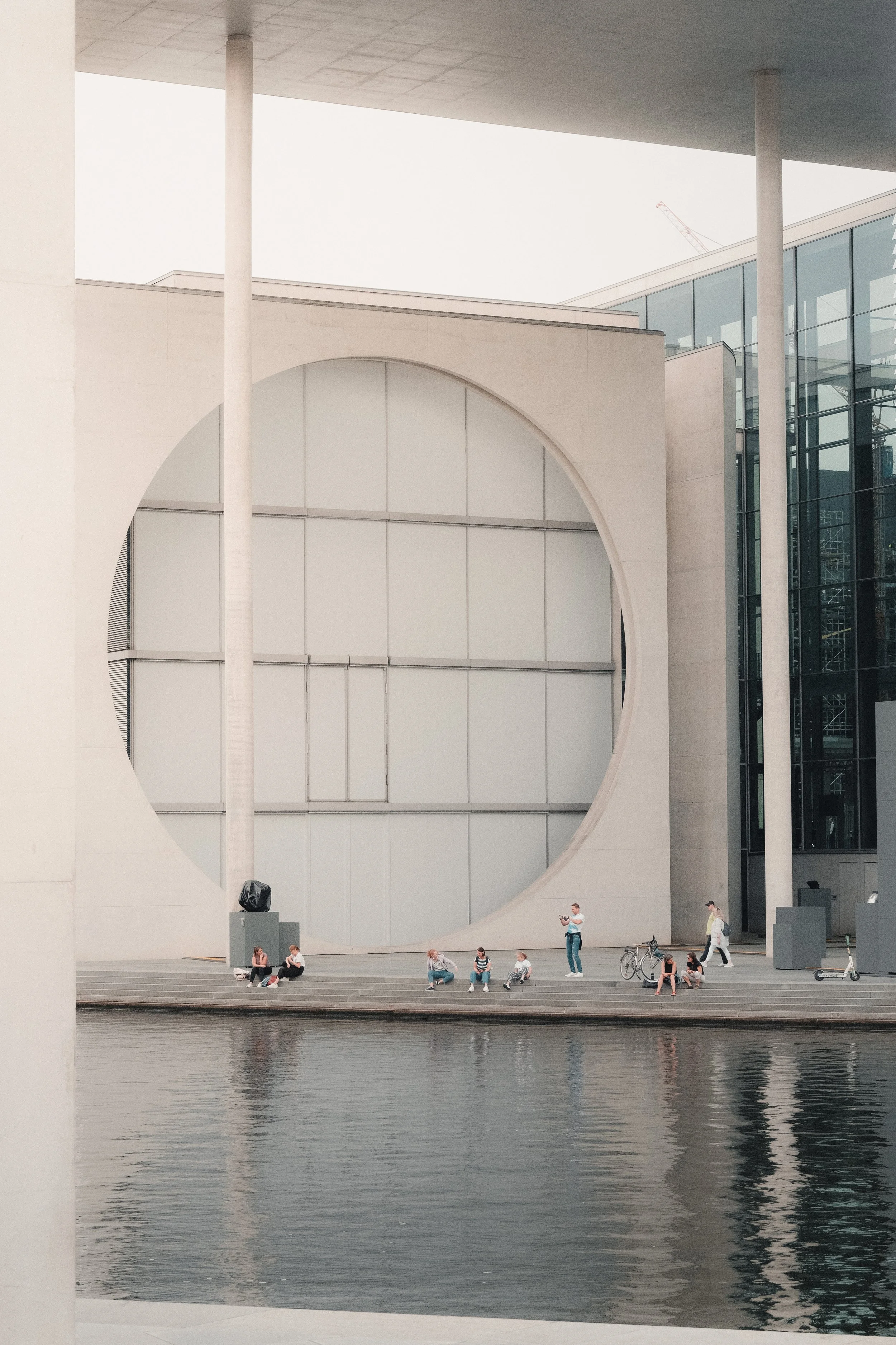 Modern gebouw met grote ronde opening, mensen zitten en staan langs het water, een fiets en kinderwagen, water reflecteert gevels