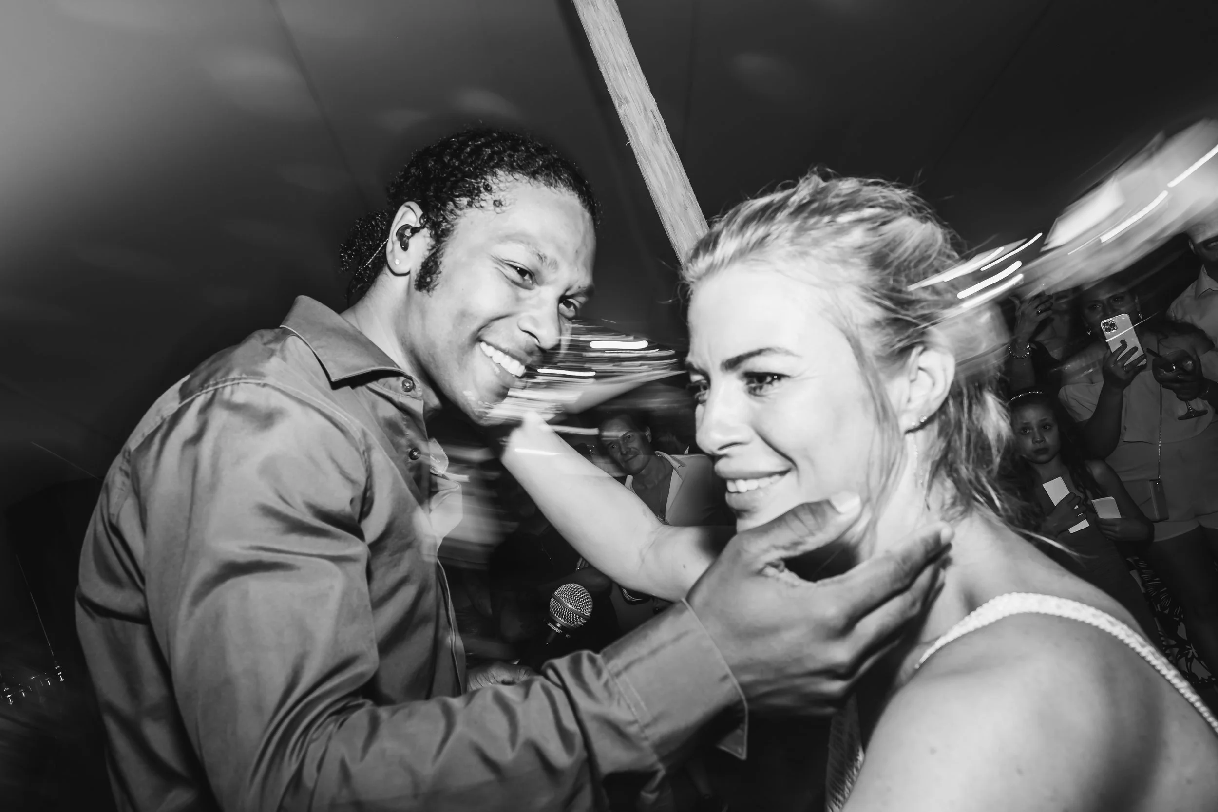 Twee mensen dansen dicht bij elkaar op een feestje, lachend. Een man met krullend haar en een vrouw met golvend haar. Op de achtergrond mensen die foto's maken en genieten.