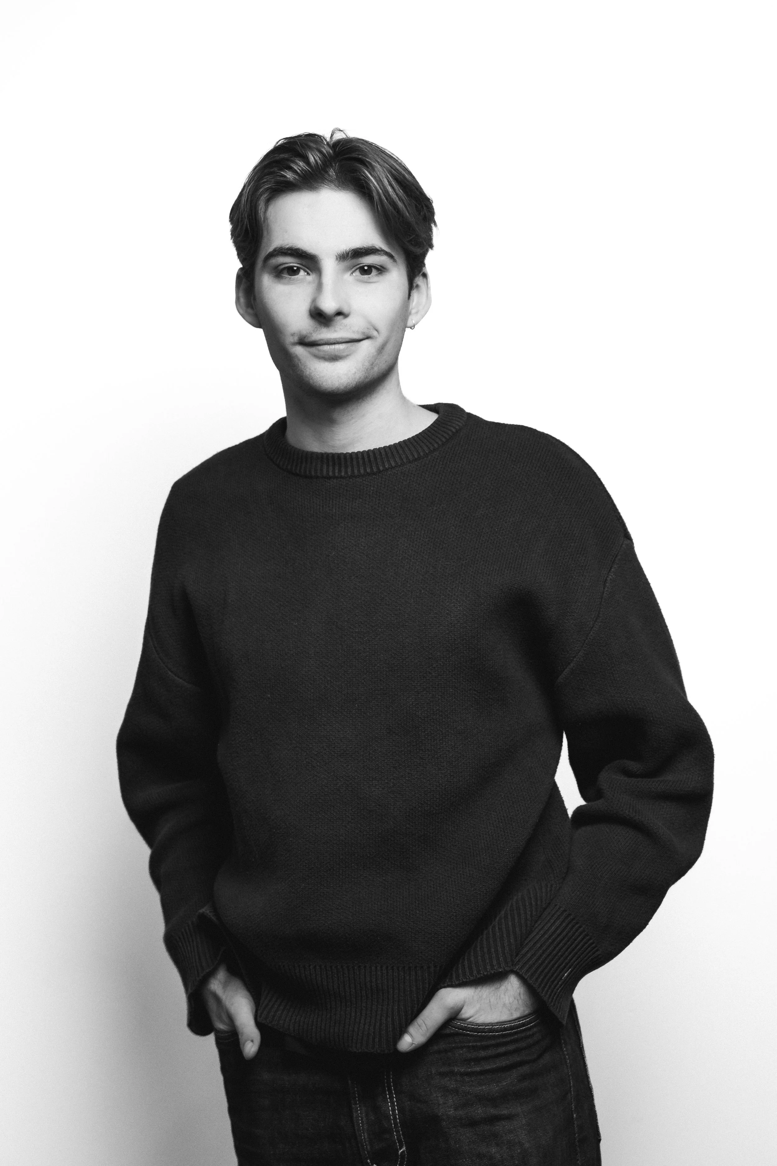 Zwart-wit foto van een jonge man met kort haar, in een zwarte pullover, die met één hand in zijn broekzak zit en in de camera kijkt met een lichte glimlach.