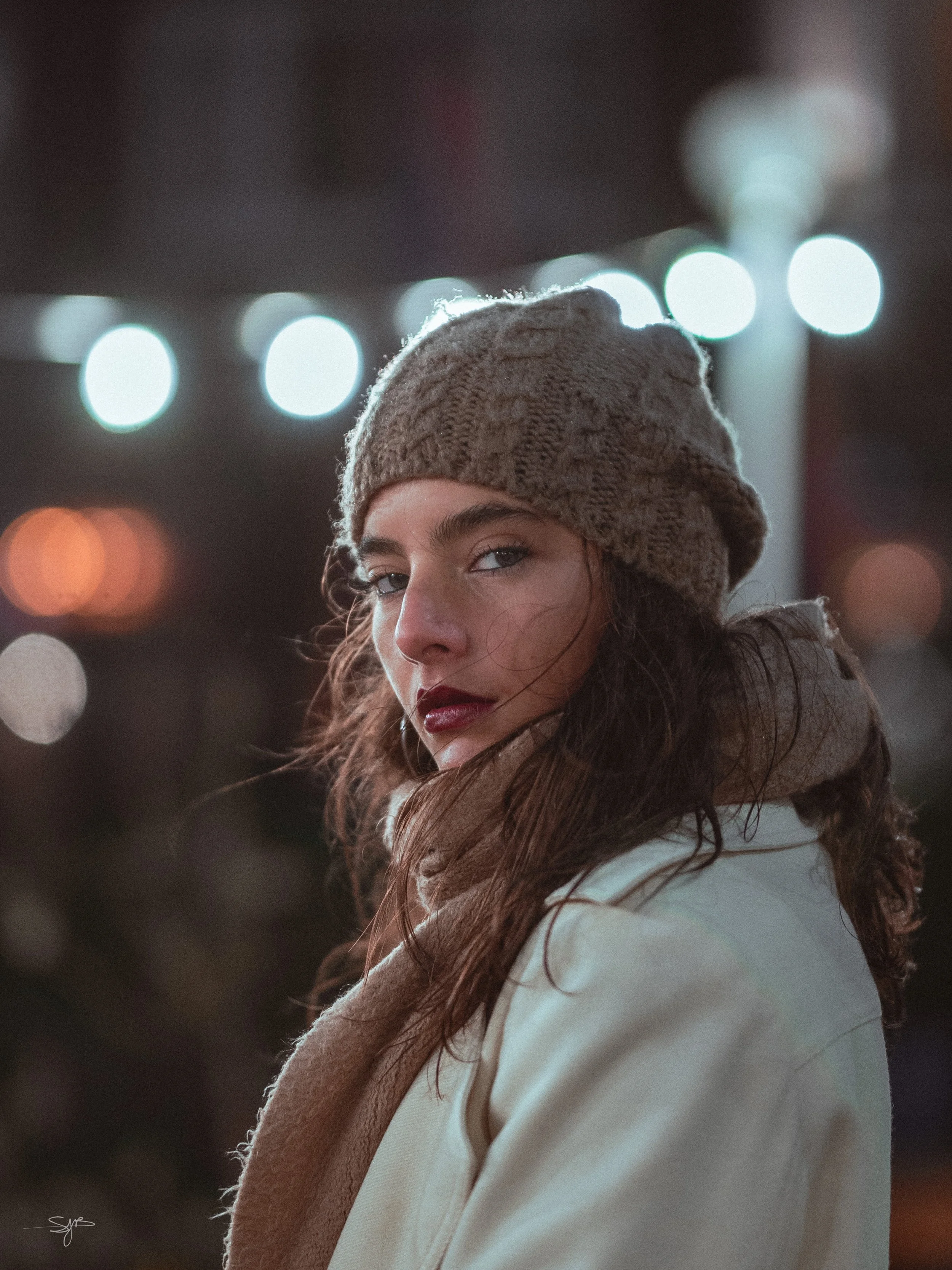 Close-up van een vrouw met lange, golvende haard, die een beige gebreide muts en een beige sjaal draagt, met een scheve blik en een paarse lippen, met een achtergrond van bokeh-light.