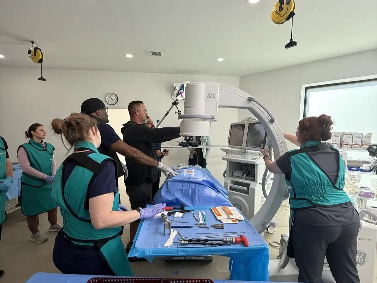 Medical education training around a surgical table for OR simulation training with a large c-arm imaging machine, likely in a cadaver lab setting.