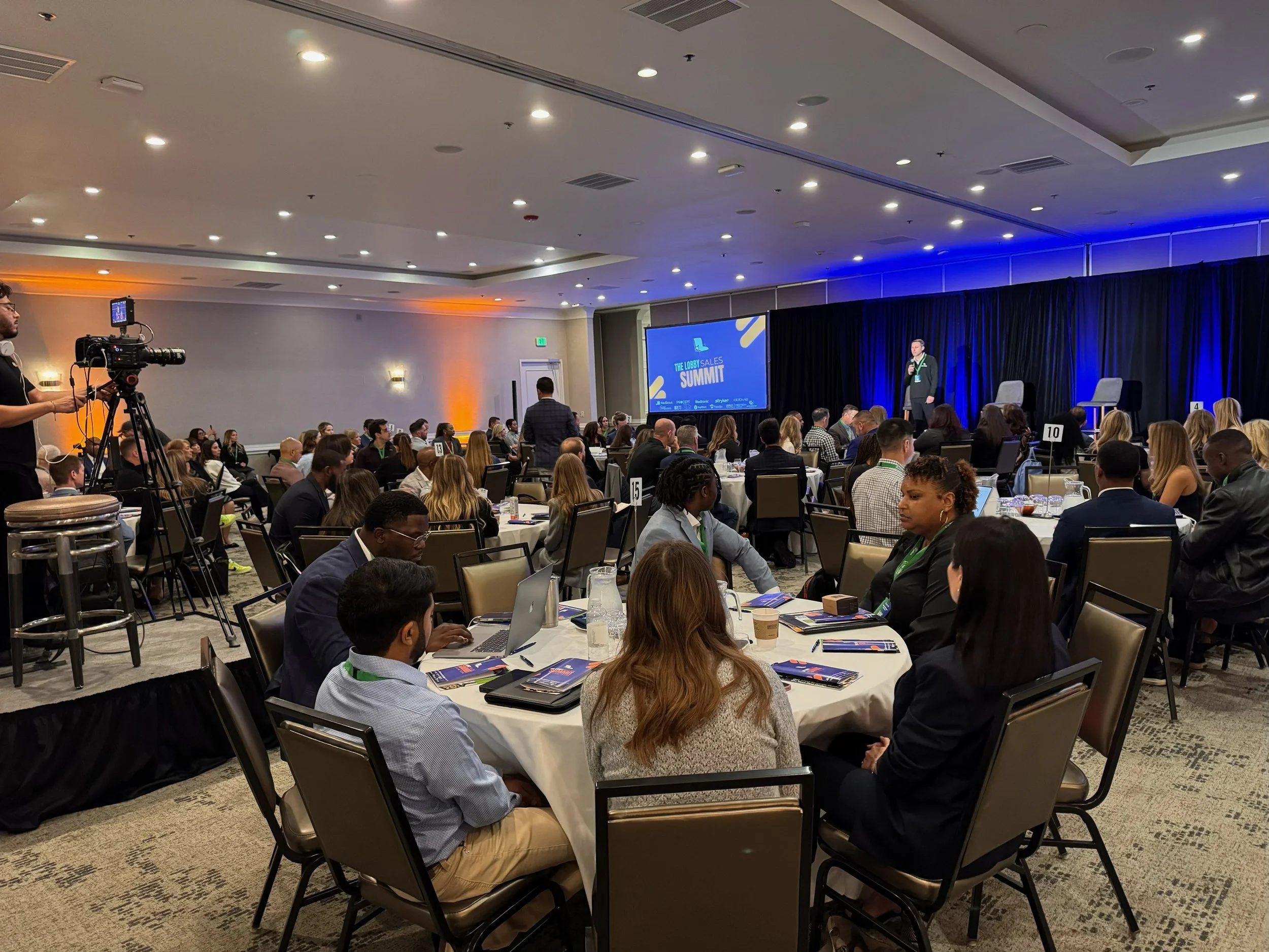 Conference room filled with attendees listening to a speaker on stage, with a large screen displaying 'The Lobby Sales Summit.' A cameraman films the event.