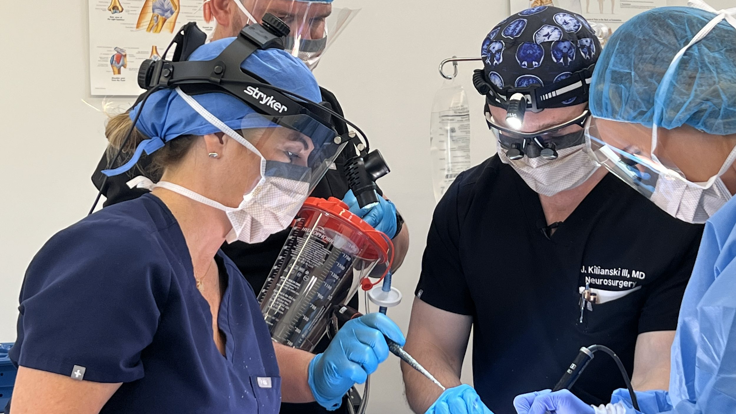 Medical team of surgeons performing a surgical procedure in METI Dallas cadaver lab OR mock room, wearing scrubs, masks, and protective headgear, with medical instruments in hand.